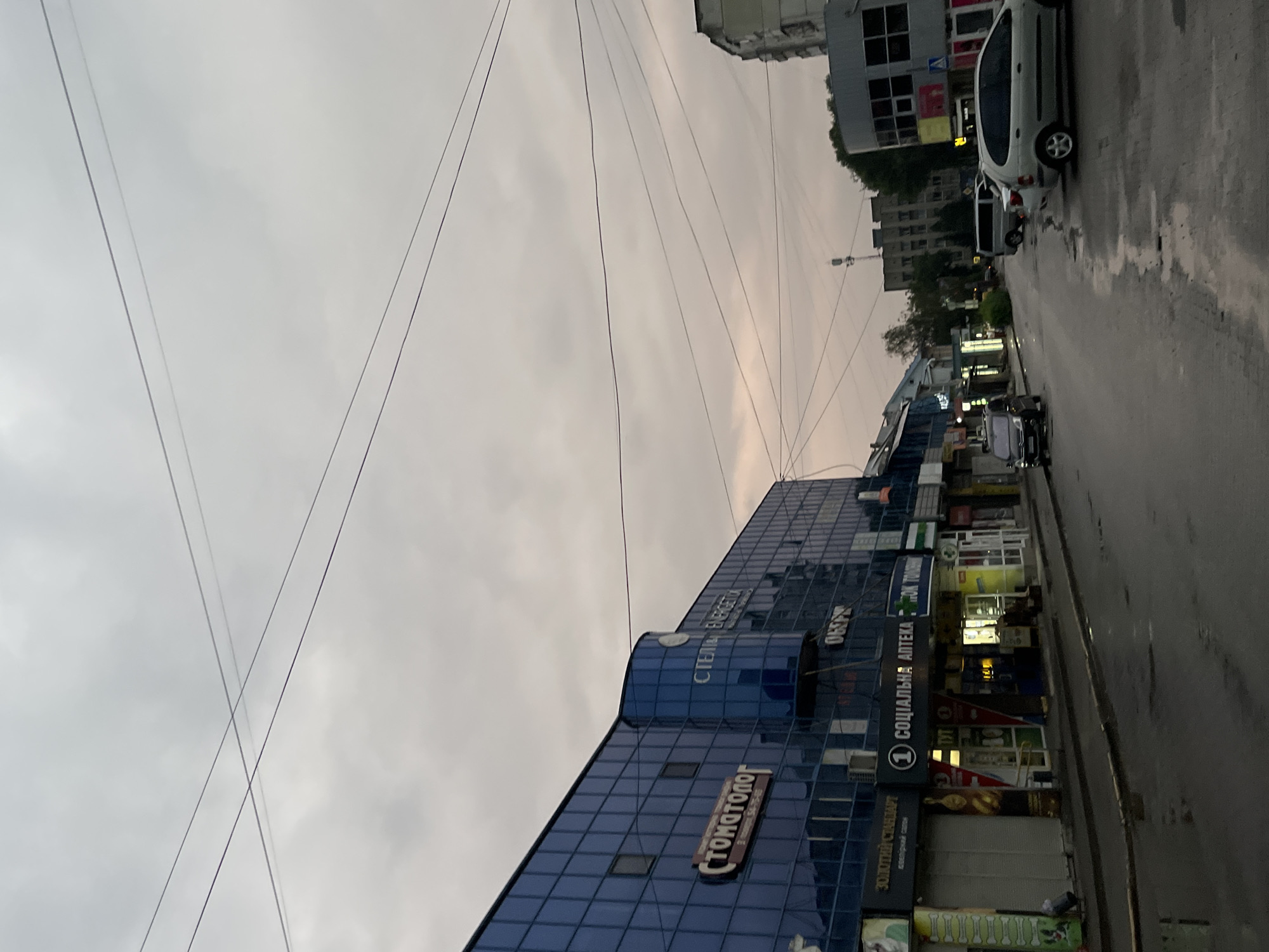 The image captures a street scene in Chernivtsi, Ukraine, under an overcast sky. In the foreground, a wet asphalt road reflects the muted light. A row of shops lines the left side of the street, with the prominent blue-glass facade of a modern building featuring signage for "СТОМАТОЛОГ" (Dentist) and other businesses. To the right, a street continues into the distance, with parked cars and more buildings visible. The overall atmosphere is subdued, suggesting it might be early morning, late evening, or a cloudy day.