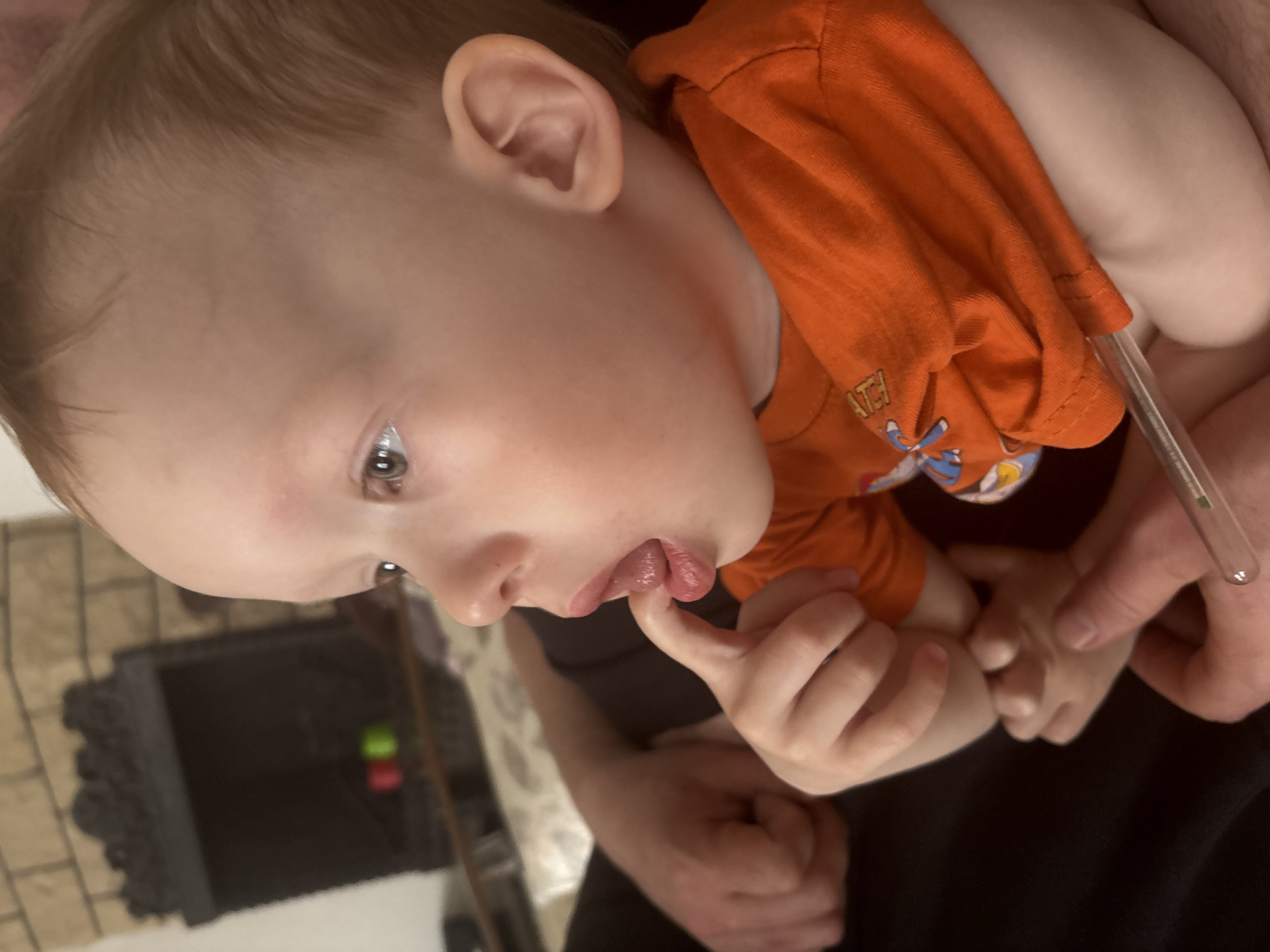 This portrait-style image, taken from a slightly high angle and rotated, captures a young child held by an adult indoors.
**Subjects:** The central subject is a fair-skinned toddler or baby with light brown hair and brown eyes, looking downwards and to the left of the frame. An adult's hands and arms are visible, holding the child.
**Activity or event:** The child is being held securely. Their left hand is raised, with an index finger near or in their mouth, and their tongue is slightly extended, suggesting self-soothing, teething, or exploration. Simultaneously, an adult's hand is holding a thin, clear glass tube, likely a thermometer, against the child's body near their armpit or side, indicating a health check or temperature measurement.
**Notable details:** The child is wearing a bright orange short-sleeved t-shirt with a faded graphic and some illegible text, possibly "ATH" as part of a larger word, on the front. The adult's clothing appears to be dark, possibly black. The background is out of focus but suggests an indoor setting, possibly a home. A dark, ornate structure resembling a fireplace or hearth is visible on the left, along with what might be a tiled floor. Some blurry red and green objects are also faintly discernible in the distance. The child's expression is one of concentration or mild curiosity rather than distress. No specific location cues for Ferma, Russia, or time of day are visible within the image.