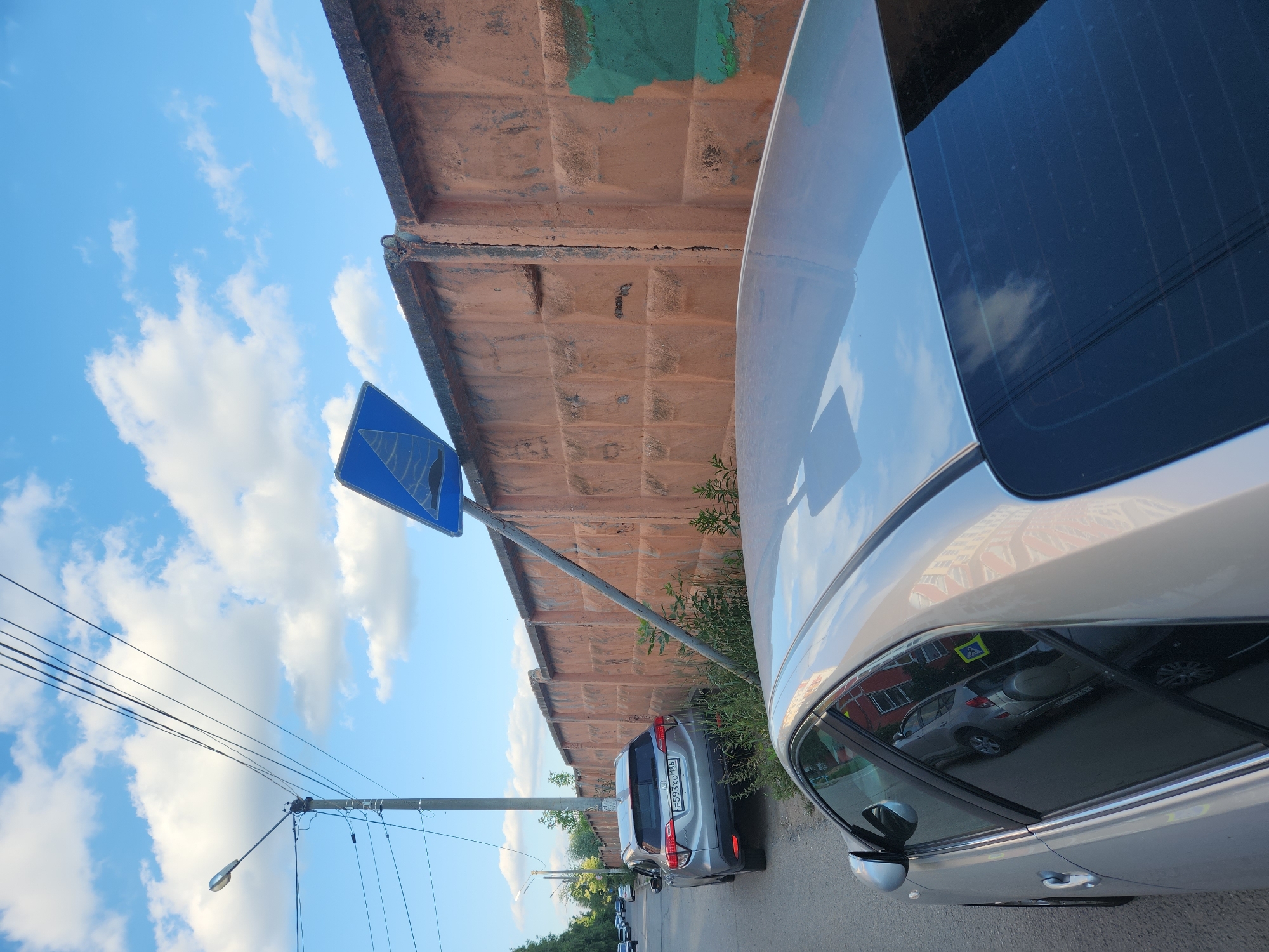 A silver car is parked next to a concrete wall under a blue sky with white clouds. The car's interior is reflected in the window, showing another car. A road sign with a blue background and a white symbol is visible on a metal pole. Power lines and a street lamp are also present. The scene appears to be in Vniissok, Russia. The time of day is likely daytime, and the weather seems clear. The license plate of a silver SUV visible in the distance reads "E593XO 186".