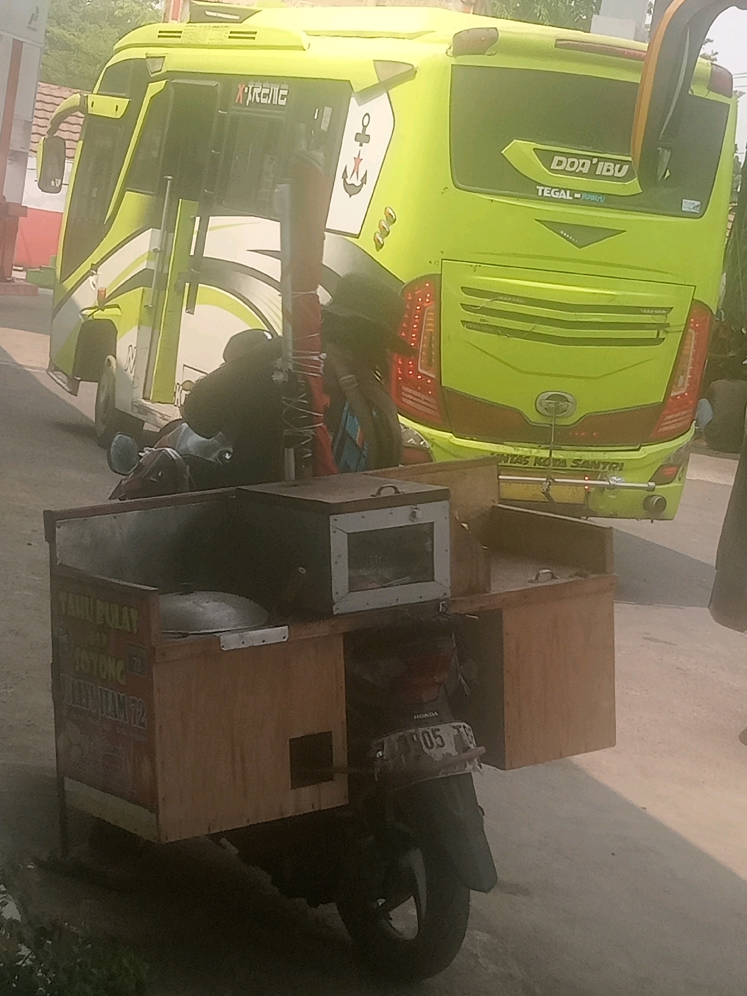 In Lebaksiu, Indonesia, a street scene unfolds with a bright yellow bus as the dominant element, partially obscured by a food cart mounted on a motorcycle. The bus, adorned with "XTREME" and "DOA' IBU" branding, appears to be parked, its rear visible with red taillights and a "TEGAL-JATENG" inscription. The food cart, made of wood, is attached to the rear of a black motorcycle with the license plate "B 205 TB". The cart contains a metal container and a small display case with glass. A person is partially visible, seated on the motorcycle, wearing dark clothing. The environment suggests an outdoor setting, possibly a roadside or parking area, with a paved surface. The overall lighting indicates it is daytime, with the sun casting shadows.