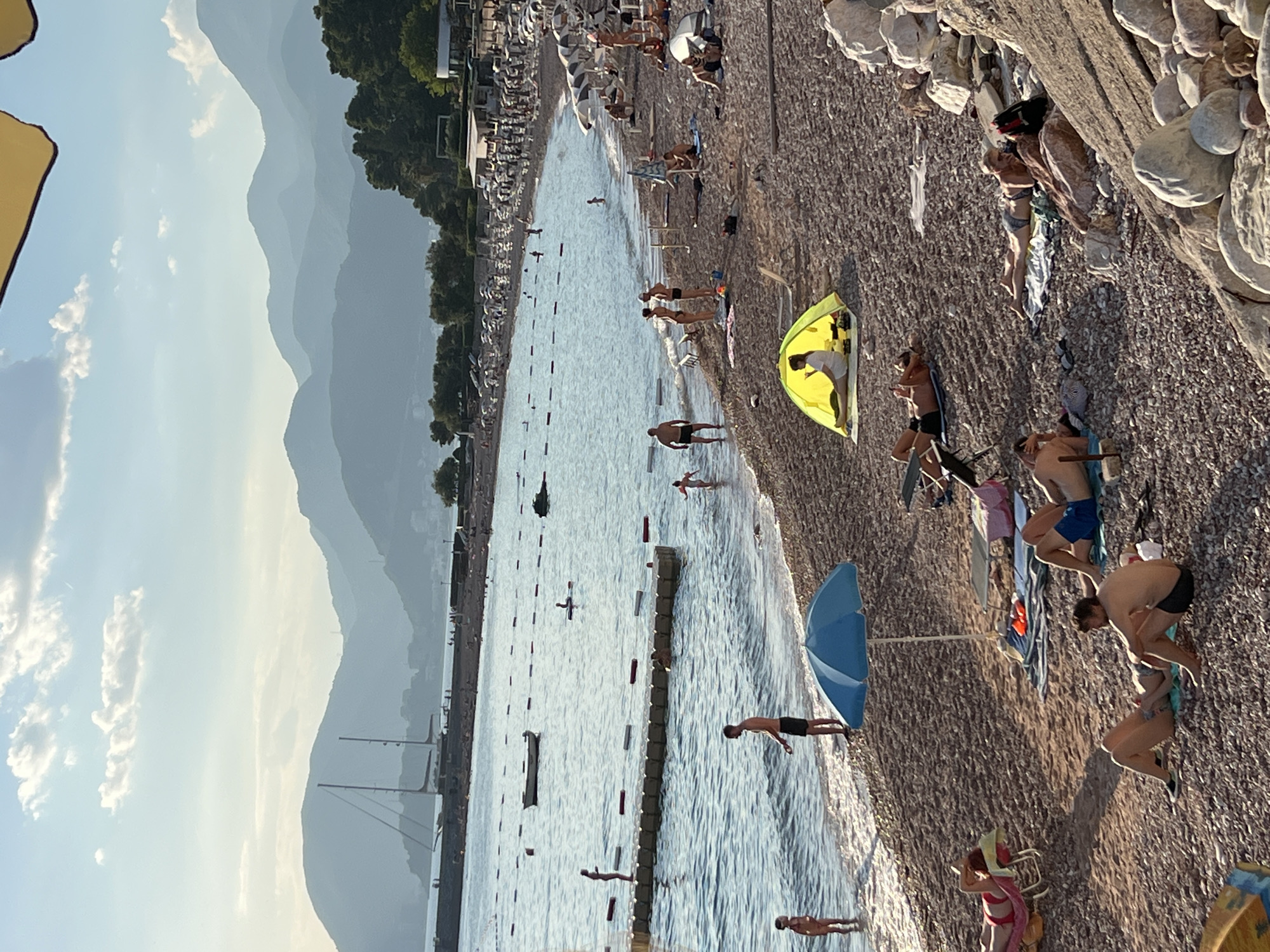 The image depicts a crowded beach scene in Sveti Stefan, Montenegro, under a bright, cloudy sky. The foreground and midground are filled with people engaged in various beach activities. Several individuals are sunbathing on towels and lounge chairs scattered across the pebbled shore. Some are lying down, others are sitting, and some appear to be chatting. A bright yellow tent is set up, with a person visible inside. A blue umbrella provides shade for another area of the beach.
The water's edge is populated with people wading and standing in the shallow sea. Further out in the water, a few individuals are swimming or engaged in water sports. A small boat is anchored in the bay, and a floating platform with people on it is visible near the shore.
In the background, a long breakwater extends into the sea, with a sailboat docked near it. Beyond the beach, the coastline is lined with trees and buildings, leading up to a series of layered, hazy mountains that dominate the horizon. The overall atmosphere is one of a lively summer day at a popular coastal resort.