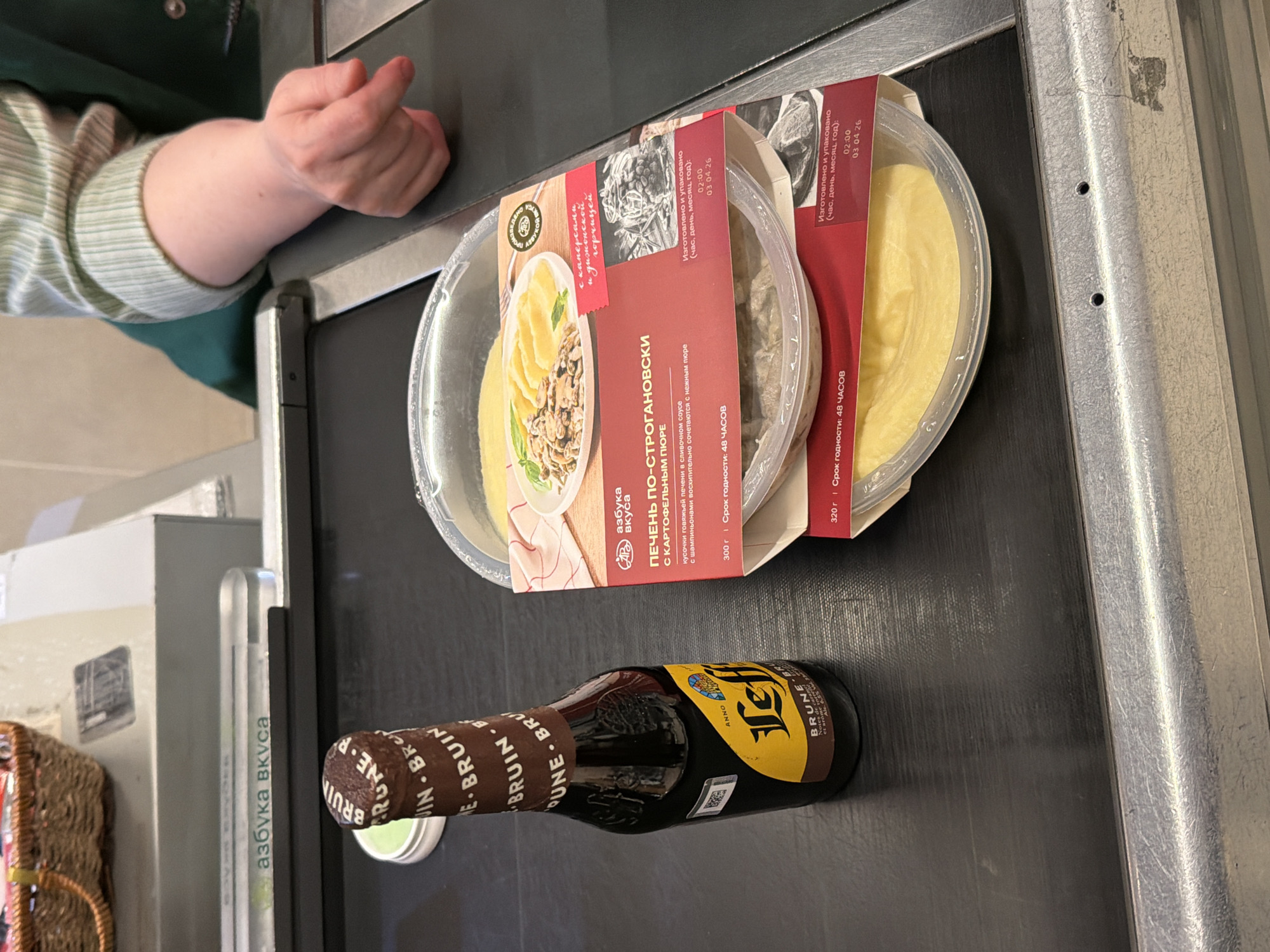 The image shows items on a checkout conveyor belt at the "Azbuka Vkusa" supermarket in Khimki, Russia. The visible items include two ready-to-eat meals, "Liver Stroganoff with Mashed Potatoes," and a bottle of Leffe Brune beer. A person's arm and hand are visible in the upper-left corner of the frame, likely belonging to the customer at the checkout. A shopping basket sits to the left of the conveyor belt. The scene suggests a customer is checking out with food and beer at the grocery store. The labels on the meals indicate they were produced and packaged on 03.04.26 at 02:00, with a shelf life of 48 hours.