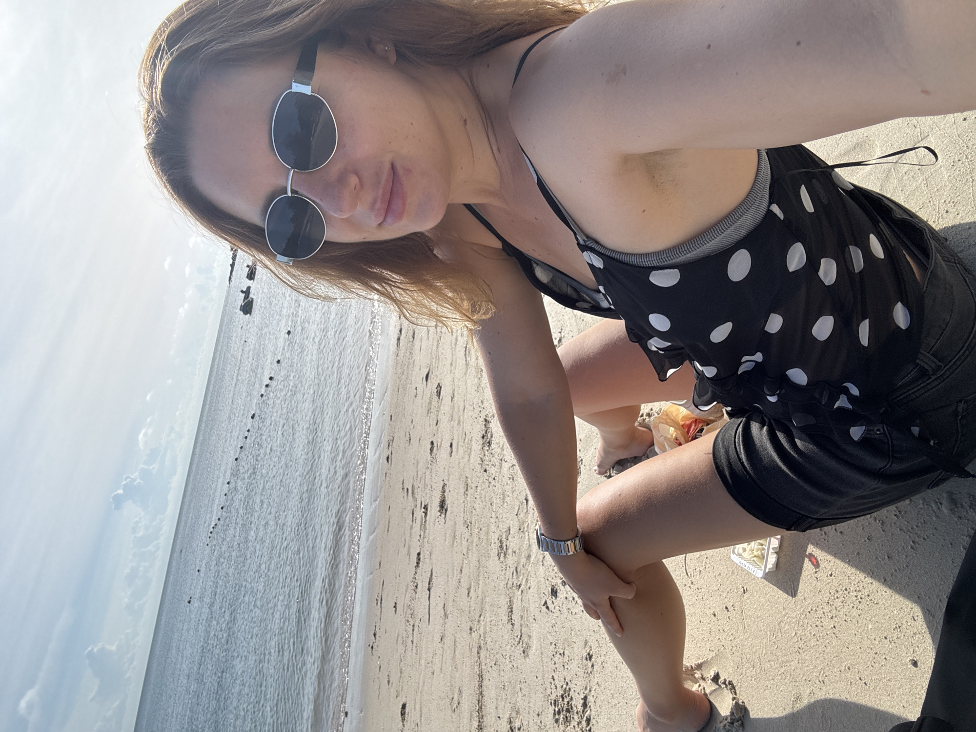 In an outdoor, full-body, selfie-style photograph, a light-skinned woman with shoulder-length, wavy brown hair and freckles on her face is seated on a sandy beach. She is wearing round, silver-rimmed sunglasses and a black polka-dot sleeveless top over a grey bra. Her legs are crossed, with her right hand resting on her left knee, where she wears a silver wristwatch. Her left leg is bent, and her foot is visible. A small white container with food is placed on the sand next to her left thigh. The background shows the ocean with a thin line of buoys and a bright, partly cloudy sky. The location is identified as Unknown, Unknown.