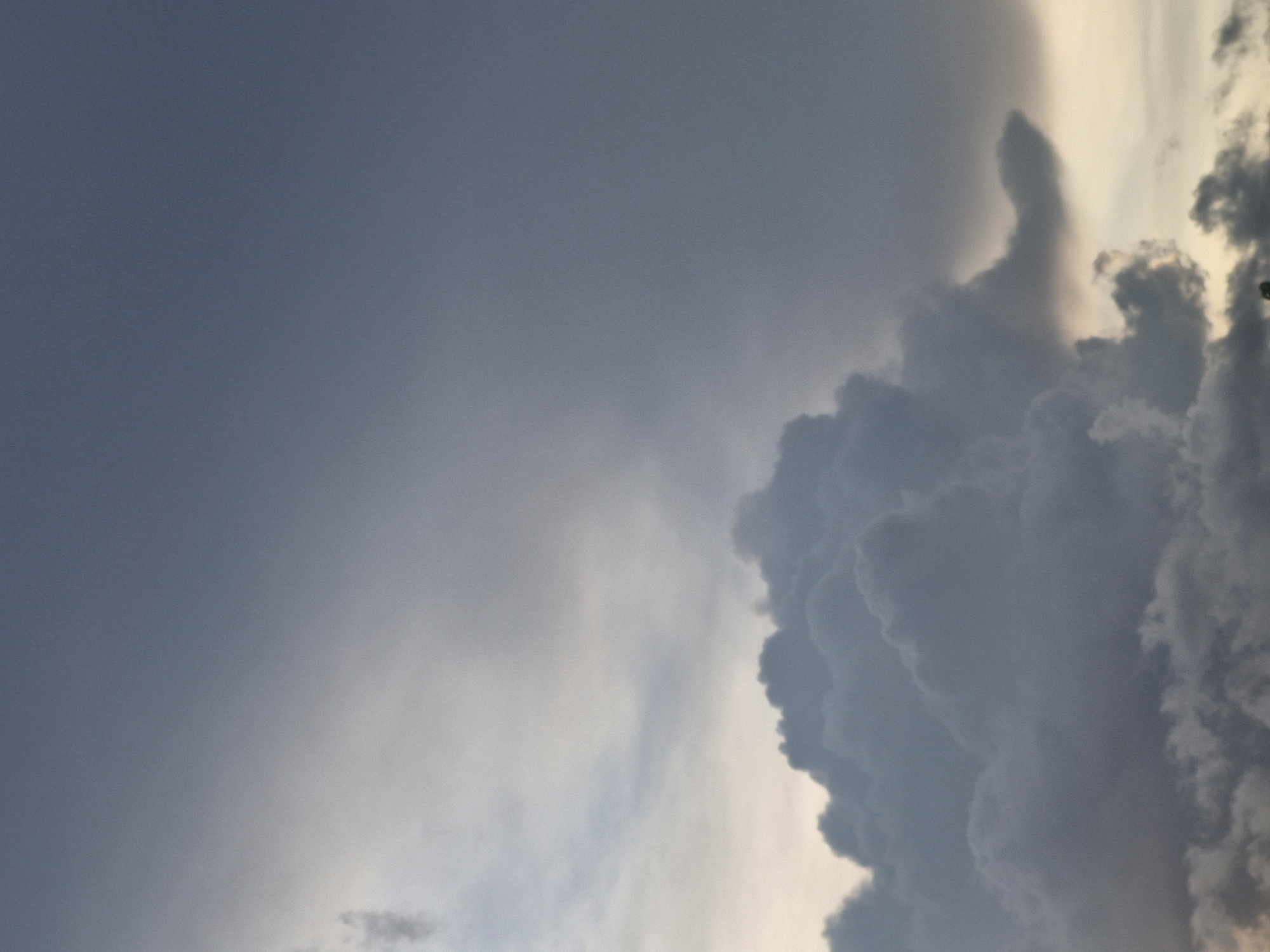 The image captures a cloudy sky over Saulgoži, Latvia. The clouds are dark and heavy, suggesting impending rain or a storm. The sky is a mix of grays and blues, with a hint of pale yellow near the horizon. There are no people, subjects, or objects visible in the frame, as the image is focused solely on the atmospheric conditions. The environment is characterized by a vast expanse of sky filled with dramatic cloud formations.