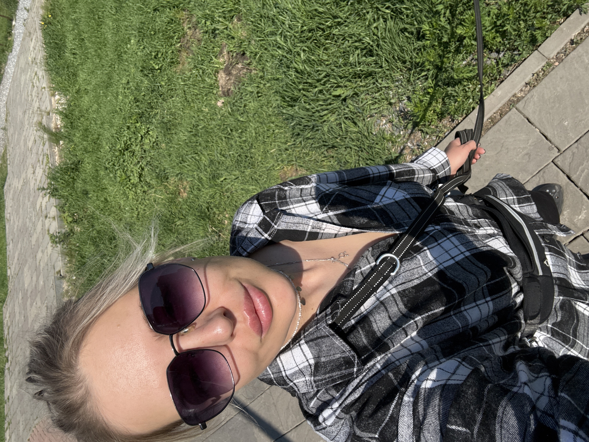 A high-angle, eye-level selfie-style photograph captures a female subject outdoors, standing on a paved surface next to a grassy area under bright sunlight. The subject, a woman with light-colored hair pulled back, wears large, dark-lensed hexagonal sunglasses with a metal frame. Her lips are prominent and appear glossy. A small piercing is visible on the upper lobe of her right ear. She is dressed in a black and white plaid long-sleeved button-up shirt, partially unbuttoned to reveal a light-colored, possibly beaded, choker necklace and a silver-colored necklace featuring a chemical structure pendant. A black strap with a metallic D-ring crosses her chest, and a black fanny pack is visible around her waist. Her right hand, in the lower right, grips a black leash extending out of frame. Her left foot, encased in a black closed-toe shoe, rests on the pavement. The immediate ground consists of light grey square pavers, separated by a concrete curb from an area of green grass and weeds to the right and in the background. Scattered patches of bare earth are visible within the grassy section. Further back, additional paved surfaces are discernible. The lighting indicates a clear, sunny day. No identifiable landmarks are present to confirm the specific location as Krasnoyarsk, Russia.