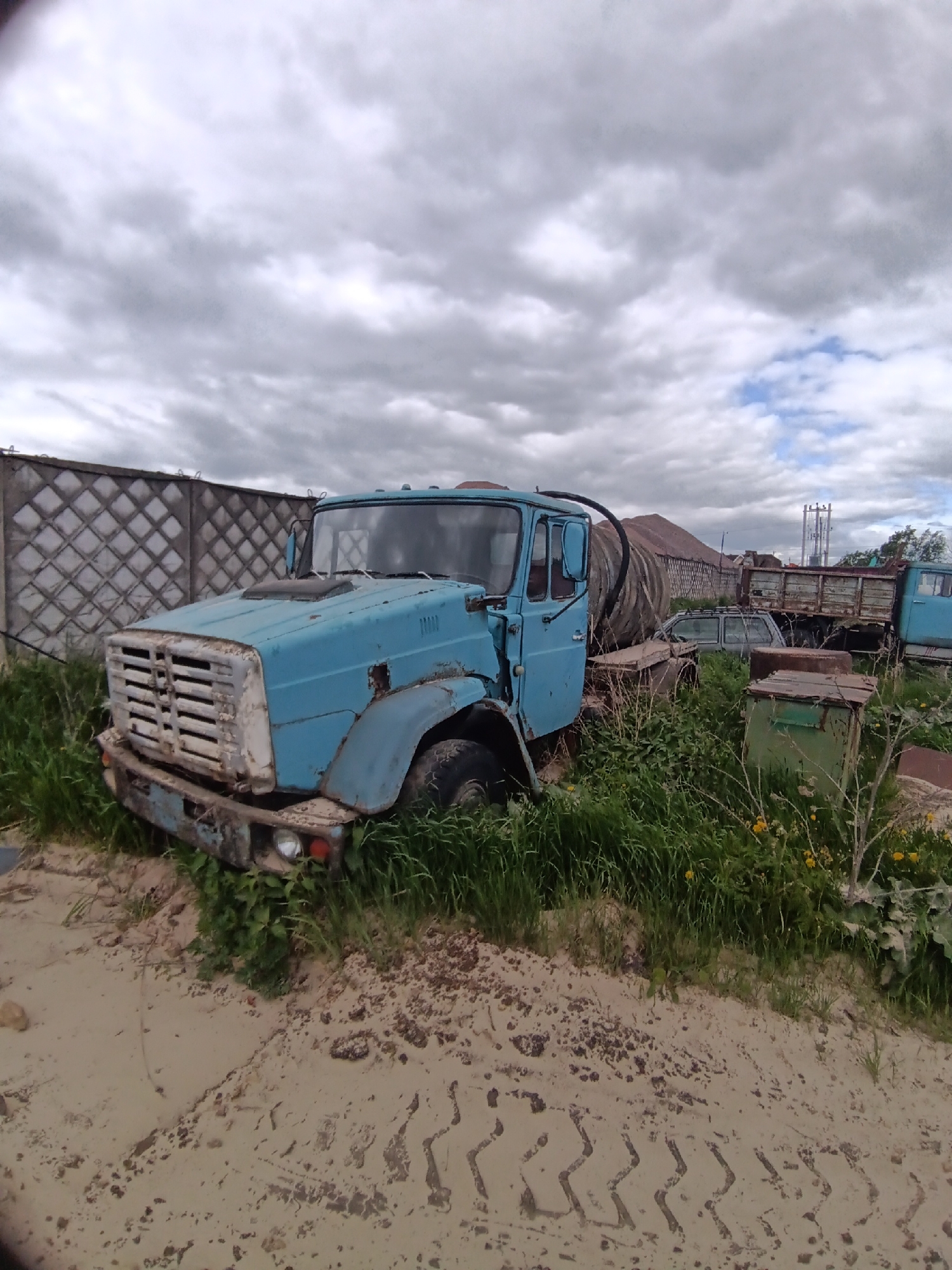 The image depicts a weathered, light blue truck partially submerged in tall grass and sand in an open area in Яльгелево, Russia. The truck's front end, including the grille and bumper, are visible, and the driver's side door is partially open. The vehicle appears to be abandoned or not in use. Other vehicles, including another truck with a flatbed, are visible in the background. A concrete block wall is visible behind the front truck. The sky is overcast with cloudy conditions. The ground is covered in a mix of sand, grass, and tire tracks.