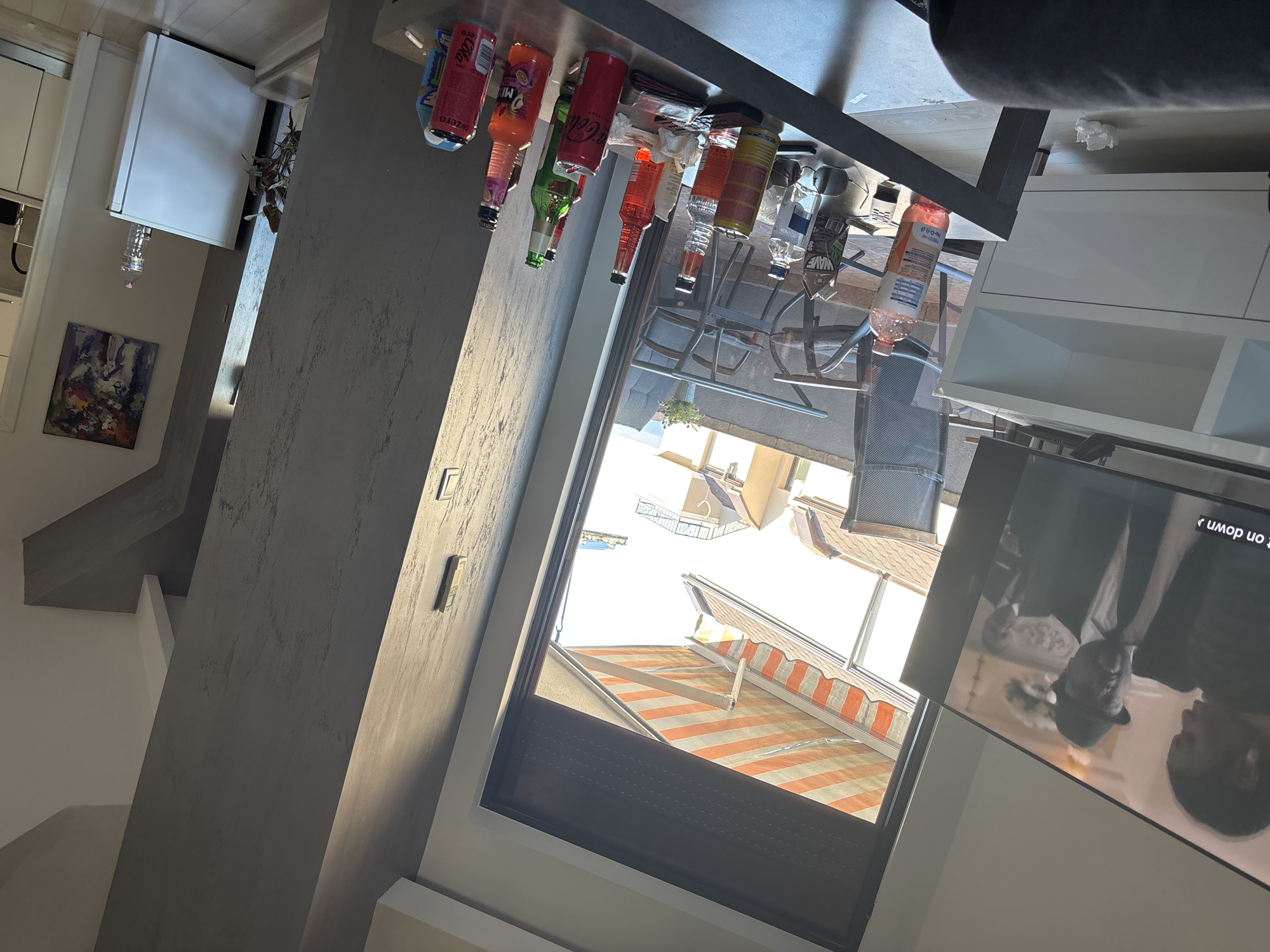 The image depicts an interior view looking out a window. Various beverages are placed on a ledge above. Through the window, a view of an outdoor patio with furniture and a striped awning is visible. Buildings are seen in the background, suggesting an urban or suburban setting. A television is visible in the foreground, displaying an image of people. The scene appears to be within a residence or establishment located in Plovdiv, Bulgaria.