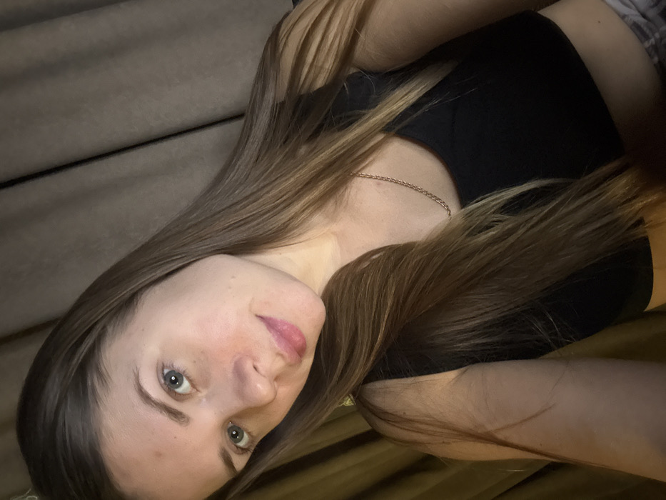 A woman with long, light brown hair is seen close-up, lying on a bed or couch. She has fair skin, green eyes, and light pink lips. She wears a black top. A thin gold necklace is visible. The background appears to be a beige or neutral-toned cushion or blanket. The lighting creates shadows across her face and hair, emphasizing the relaxed setting.
