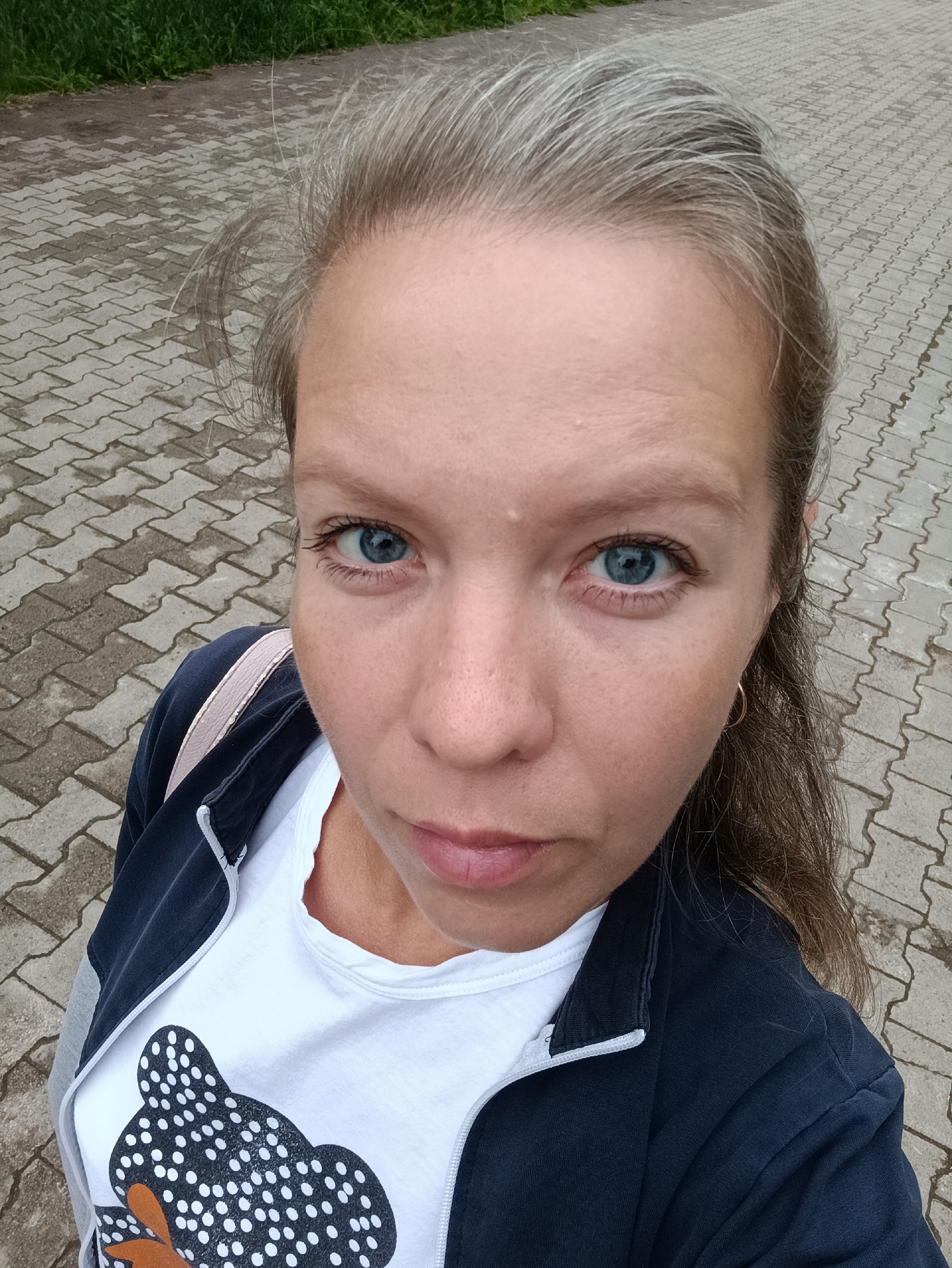 An outdoor, high-angle, close-up selfie features a woman with fair skin, light brown hair with grey streaks, and blue eyes. She is wearing a dark blue zip-up jacket over a white t-shirt with a black and white polka dot bear graphic. The background consists of a paved walkway made of interlocking grey concrete pavers, with a strip of green grass visible in the upper left corner. The location is identified as Chaikovsky, Russia.
