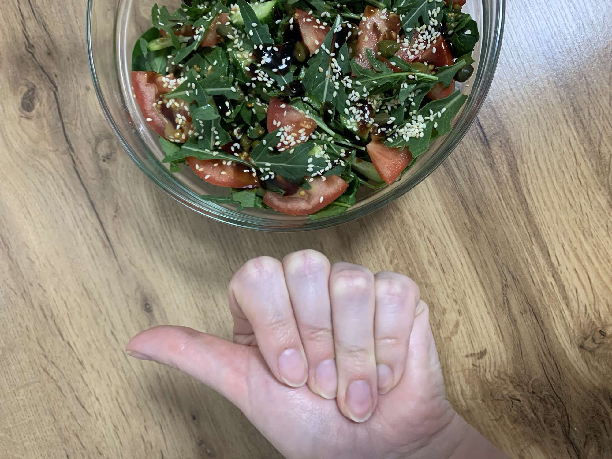An overhead shot captures a human hand making a "thumbs up" gesture positioned in the foreground, with a vibrant salad in a clear glass bowl occupying the background. The scene is set on a light brown wooden surface with visible grain patterns, likely an indoor table or counter in Ternopil, Ukraine.
The hand, belonging to an adult with light skin and natural, unpolished fingernails, extends its thumb upward as a sign of approval or satisfaction. The fingers are curled into a fist.
The salad in the glass bowl appears fresh and appealing. It primarily consists of dark green arugula leaves, interspersed with bright red, thinly sliced tomatoes and dark black olives. Green capers and a generous sprinkling of white sesame seeds are distributed throughout the salad, adding texture and color. A dark, glossy balsamic glaze or dressing is visible drizzled over the ingredients, adding to the visual appeal. The combination of ingredients suggests a healthy and flavorful dish.
No specific time of day or weather conditions can be inferred from the image, and there is no visible text. The overall impression is one of contentment and appreciation for the food.