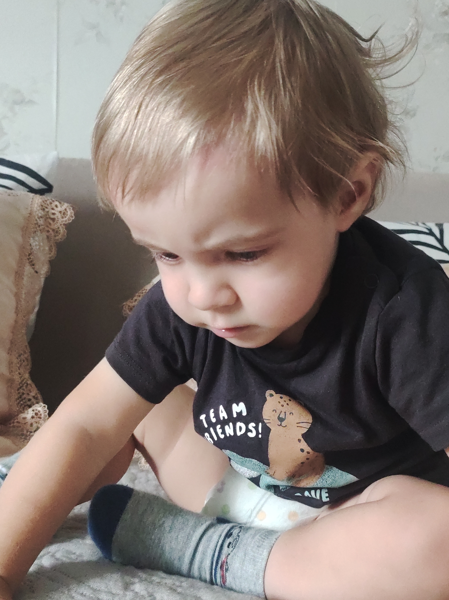A young child, likely a toddler, with light blonde hair and fair skin, is seated indoors, looking downwards with a focused or contemplative expression. The child is wearing a black short-sleeved t-shirt with a graphic design, a diaper, and one visible grey sock with a blue toe and heel. A small red mark is noticeable on the child's forehead. The left arm is extended forward and downwards, suggesting interaction with something out of frame.
The setting appears to be a cozy indoor space, possibly a bedroom or living room, with soft furnishings. To the left, a light-colored pillow with intricate lace trim is visible. The child is sitting on what looks like a light-colored, patterned blanket or bedspread. The background features a subtly patterned, light-colored wall and hints of another striped pillow or headboard.
On the child's black t-shirt, the words "TEAM FRIENDS!" are clearly visible, accompanied by a cartoon illustration of a smiling spotted big cat, resembling a leopard or cheetah, in shades of brown and light blue. The overall atmosphere is calm and quiet, with the child engrossed in an activity.