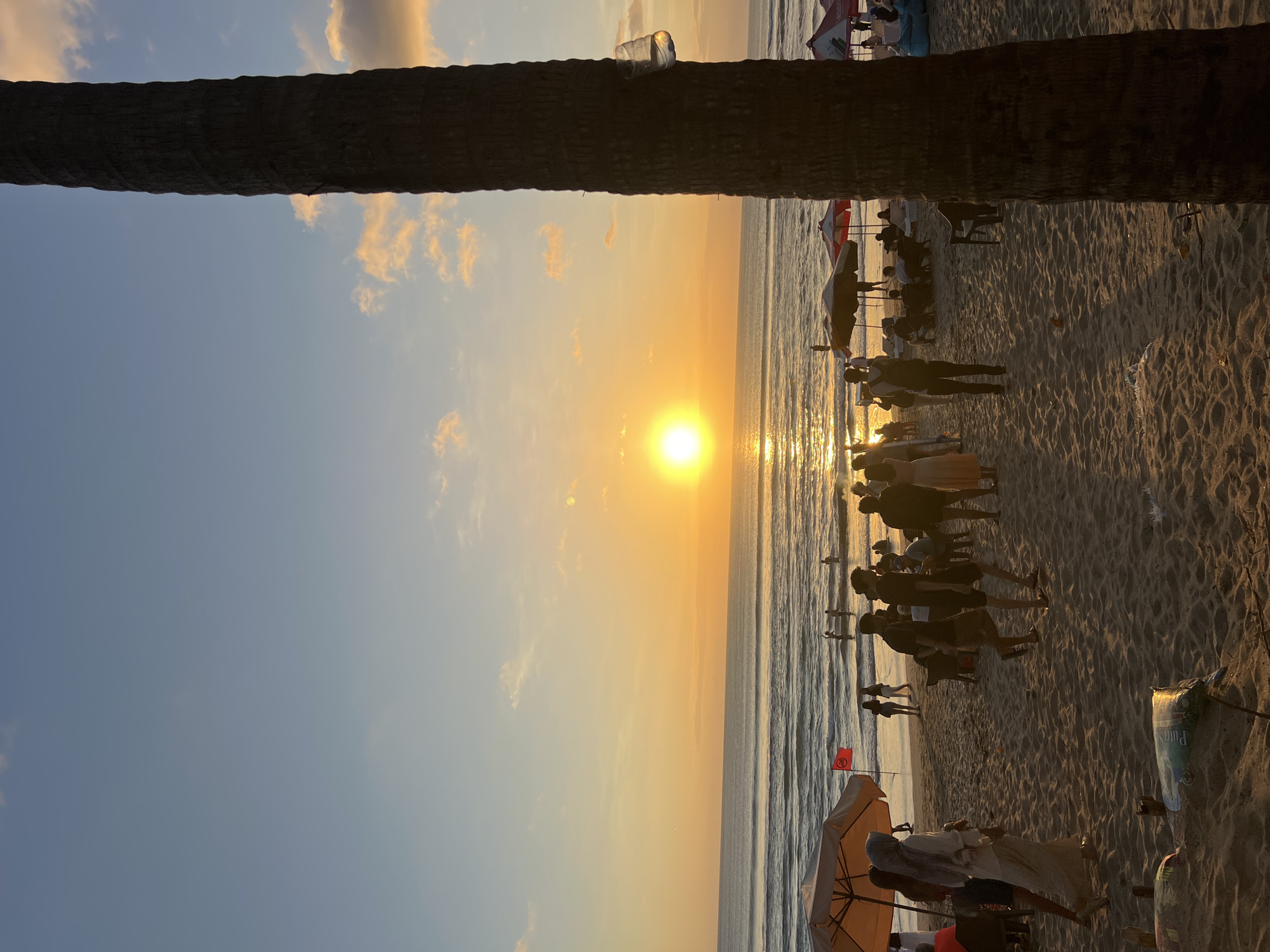 This is a photograph taken at sunset on the beach in Kuta, Bali, Indonesia. The sun is low on the horizon, casting a warm, golden glow over the scene. The sky is a gradient of blue and orange, with scattered clouds tinged with pink and yellow.
In the foreground, a large palm tree trunk dominates the right side of the frame, its rough texture visible. The sandy beach is populated with a variety of people enjoying the evening. Some are walking along the shoreline, while others are gathered under beach umbrellas, likely relaxing or socializing. There are beach chairs and some vendors' stalls visible in the midground.
The ocean stretches out to the horizon, with gentle waves rolling onto the shore. The setting sun creates a shimmering reflection on the water's surface. Several individuals are in the water, some appearing to be wading or swimming. In the distance, more beachgoers are visible, further populating the coastline.
The overall atmosphere is peaceful and serene, characteristic of a sunset on a popular beach. The image captures a moment of leisure and natural beauty in a well-known tourist destination.