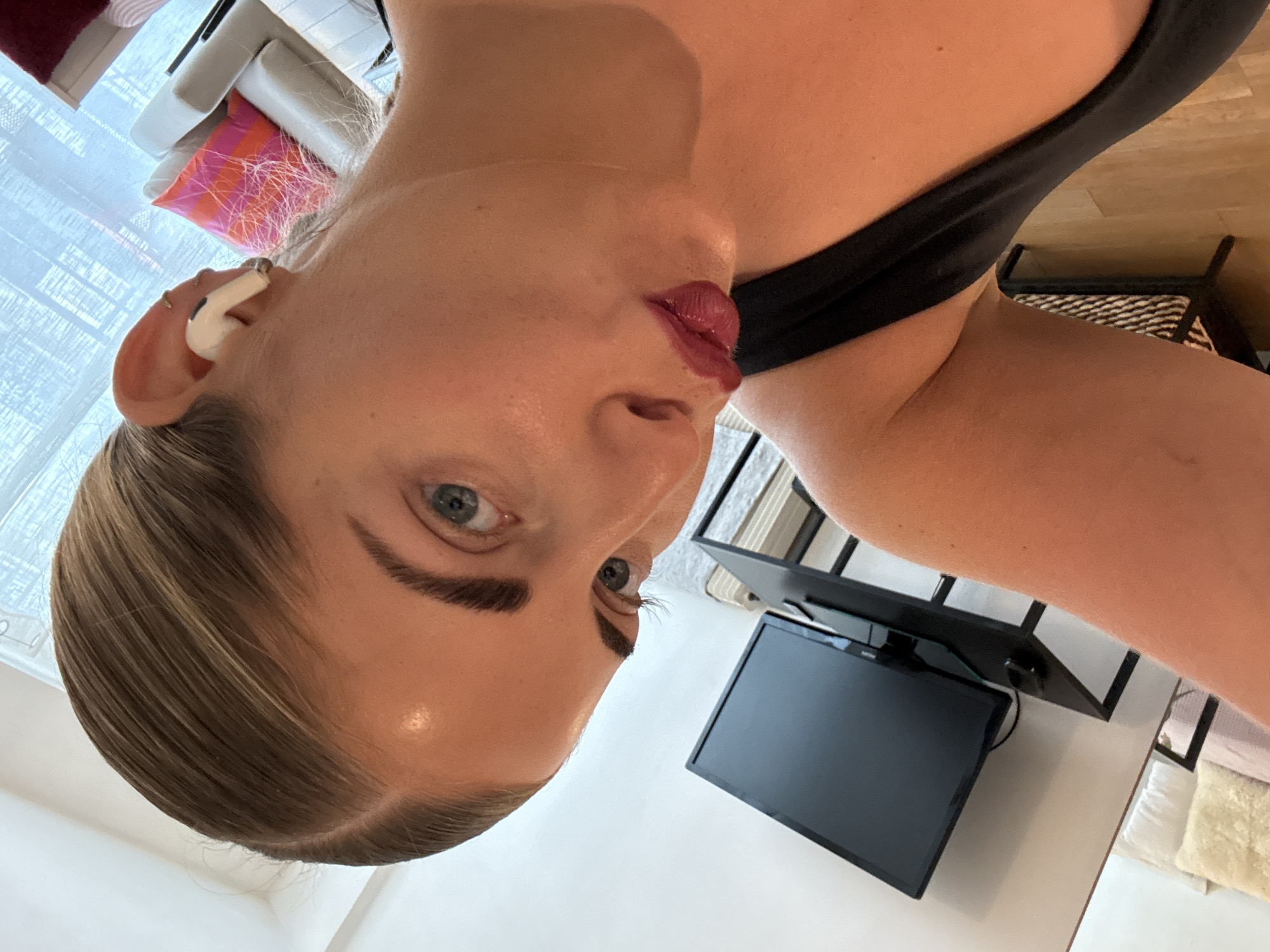 A rotated close-up shot features a blonde woman wearing an earpiece. She has light skin, blue eyes, and dark red lipstick. The background includes a television, a black shelf, a chair with a patterned cushion, and a window with a white curtain. It is impossible to definitively determine the location as Paris, France, without additional context.