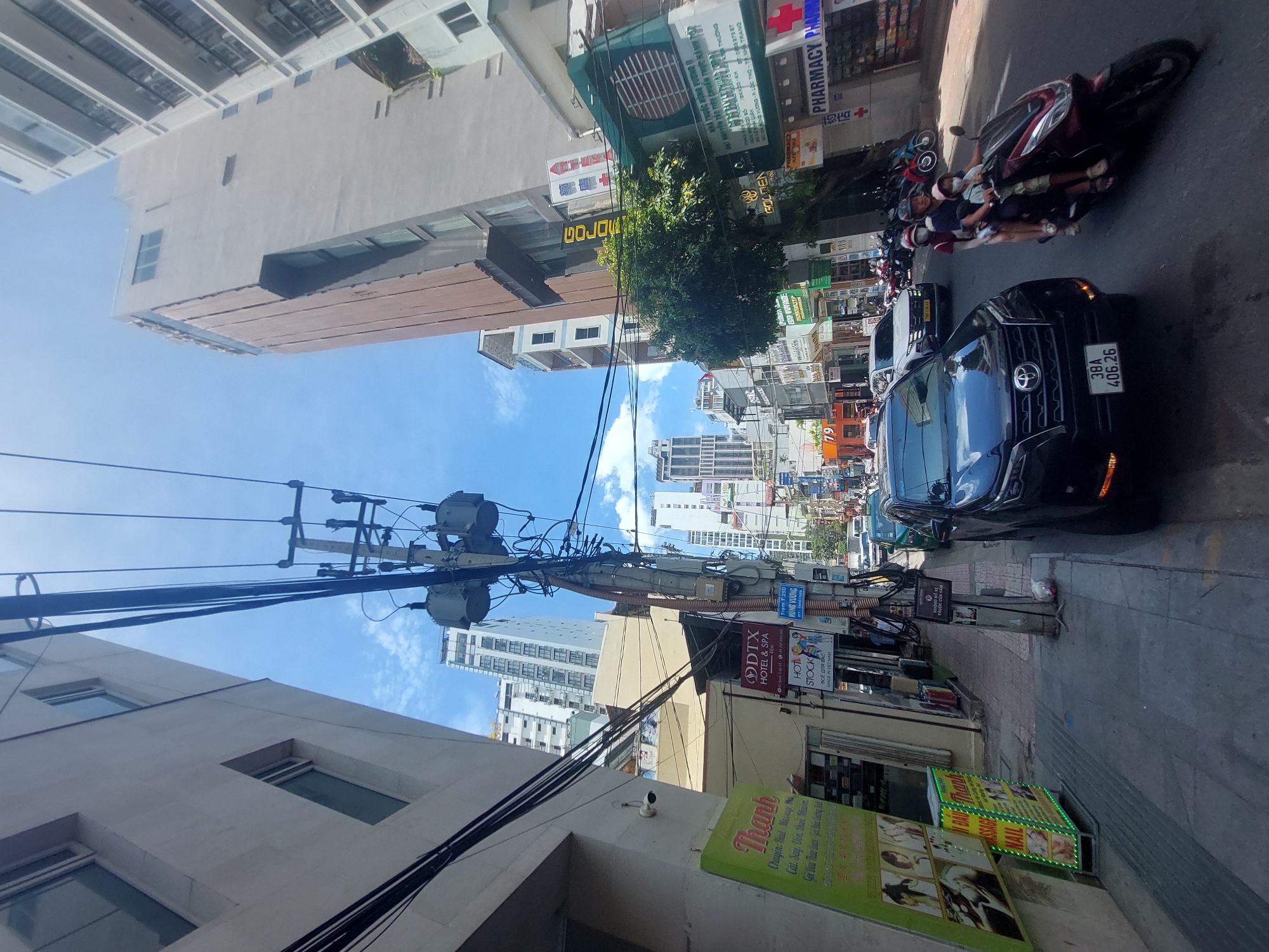 This is a street-level, forward-facing photograph taken in Nha Trang, Vietnam, under a clear sky with scattered clouds. The scene depicts a busy urban environment with a mix of modern and older buildings, including high-rises and smaller commercial establishments lining the street. Overhead, a dense network of electrical wires and utility poles crisscrosses the sky, casting shadows on the buildings and the street below.
In the foreground, a dark SUV with license plate "38A 406.26" is parked on the right side of the road. To its right, a red scooter carrying three individuals – an adult male driver and two children, presumably passengers – is traveling away from the camera. All three are wearing face masks. The scooter has a license plate that reads "TRAM 725D HUNG VUONG".
The street itself is paved with asphalt, and a sidewalk runs along the left side of the image. The sidewalk features a green illuminated sign advertising "THÀNH Massage Nail", with images of beauty treatments. Further down the sidewalk, several businesses are visible, including "DTX HOTEL & SPA", a shop with a "HOT STOCK" sign, and a "PHARMACY" store. Motorbikes are parked along the sidewalk in front of some businesses.
The architecture varies, with tall, modern buildings dominating the background. Some buildings have visible windows and balconies. The overall impression is one of a vibrant, bustling commercial district. The photograph appears to have been taken during daylight hours, likely in the mid-morning or afternoon, given the bright sunlight and clear sky.