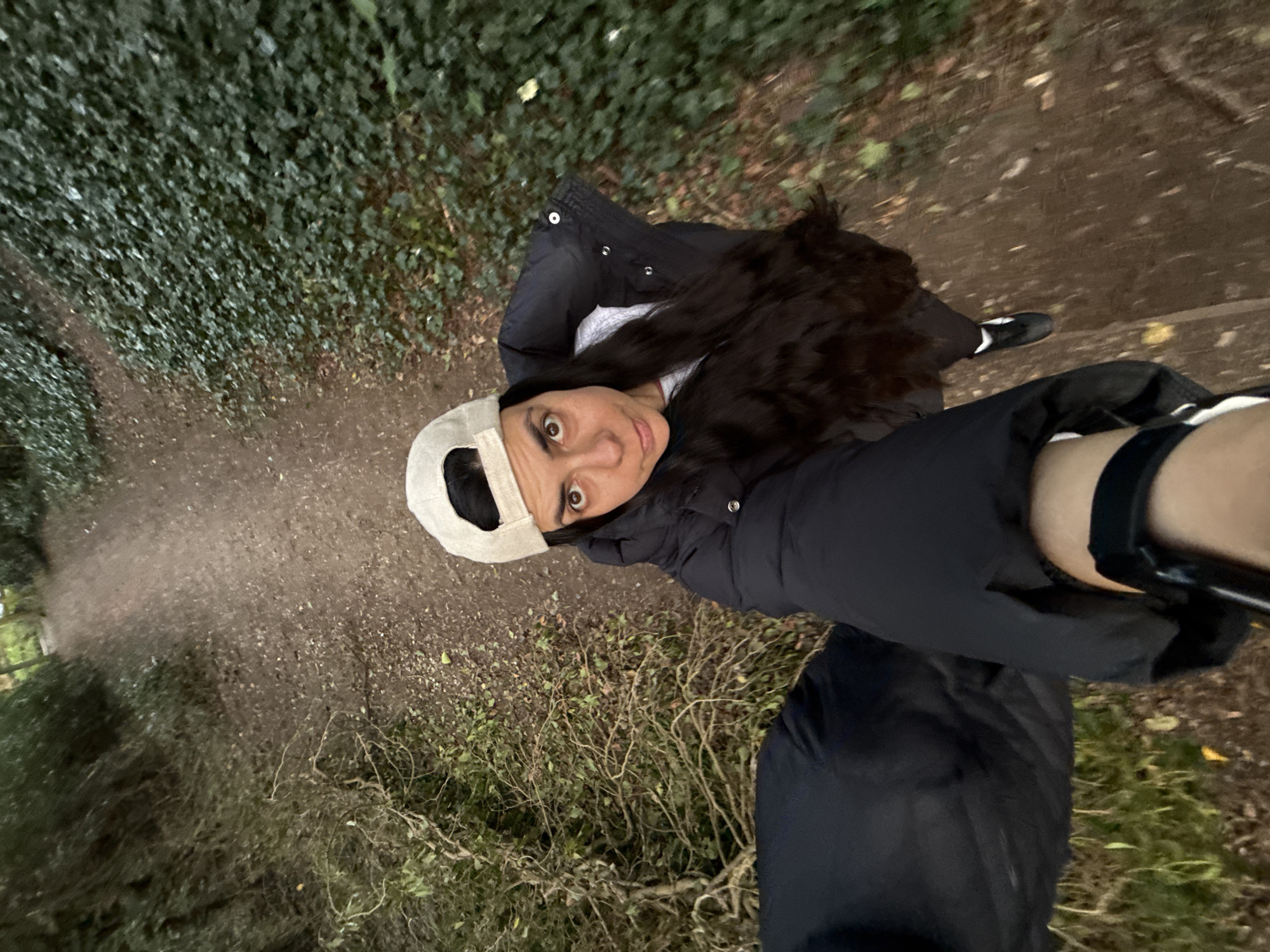 A woman wearing a black puffer jacket and a white baseball cap stands on a dirt path in what appears to be a wooded area. To her left is a dense wall of ivy or similar vegetation, while to her right are low-lying shrubs and sparse ground cover. The path is dirt or gravel. The woman is holding a camera or phone on an arm extension, capturing a selfie from a high angle. One of her feet, encased in a black and white athletic shoe, is visible.
I lack the ability to determine the specific location within Great Yarmouth.