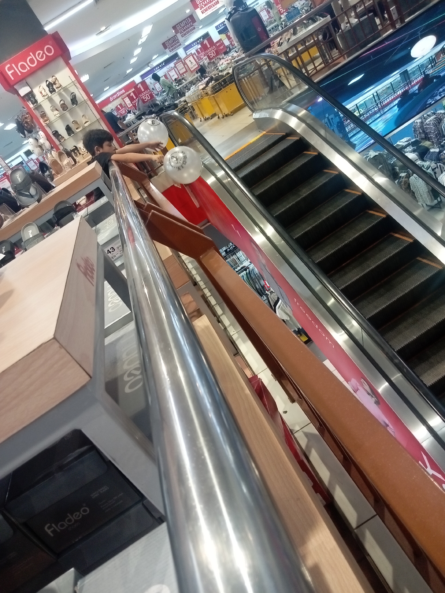 The image captures a scene inside a retail store, likely a department store or a shoe store, in Batam, Indonesia. The perspective is from a slightly elevated angle, looking down towards an escalator. In the foreground, a young boy with dark hair is leaning over a railing, reaching towards two white balloons with some branding on them.
To the left of the boy is a display stand for "Fladeo" brand shoes, showcasing various footwear. Behind the display, shelves filled with shoe boxes are visible. The overall environment appears to be well-lit with artificial lighting from the ceiling.
The escalator is a prominent feature, with its metallic handrails and dark, ribbed steps. Advertisements and promotional banners are visible along the side of the escalator, one of which reads "freedom". In the background, more merchandise, including clothing and other retail items, is displayed on shelves and racks. Other shoppers and store staff can be seen in the distance, suggesting a moderately busy commercial setting. The floor tiles and the general store layout are typical of Indonesian retail spaces.