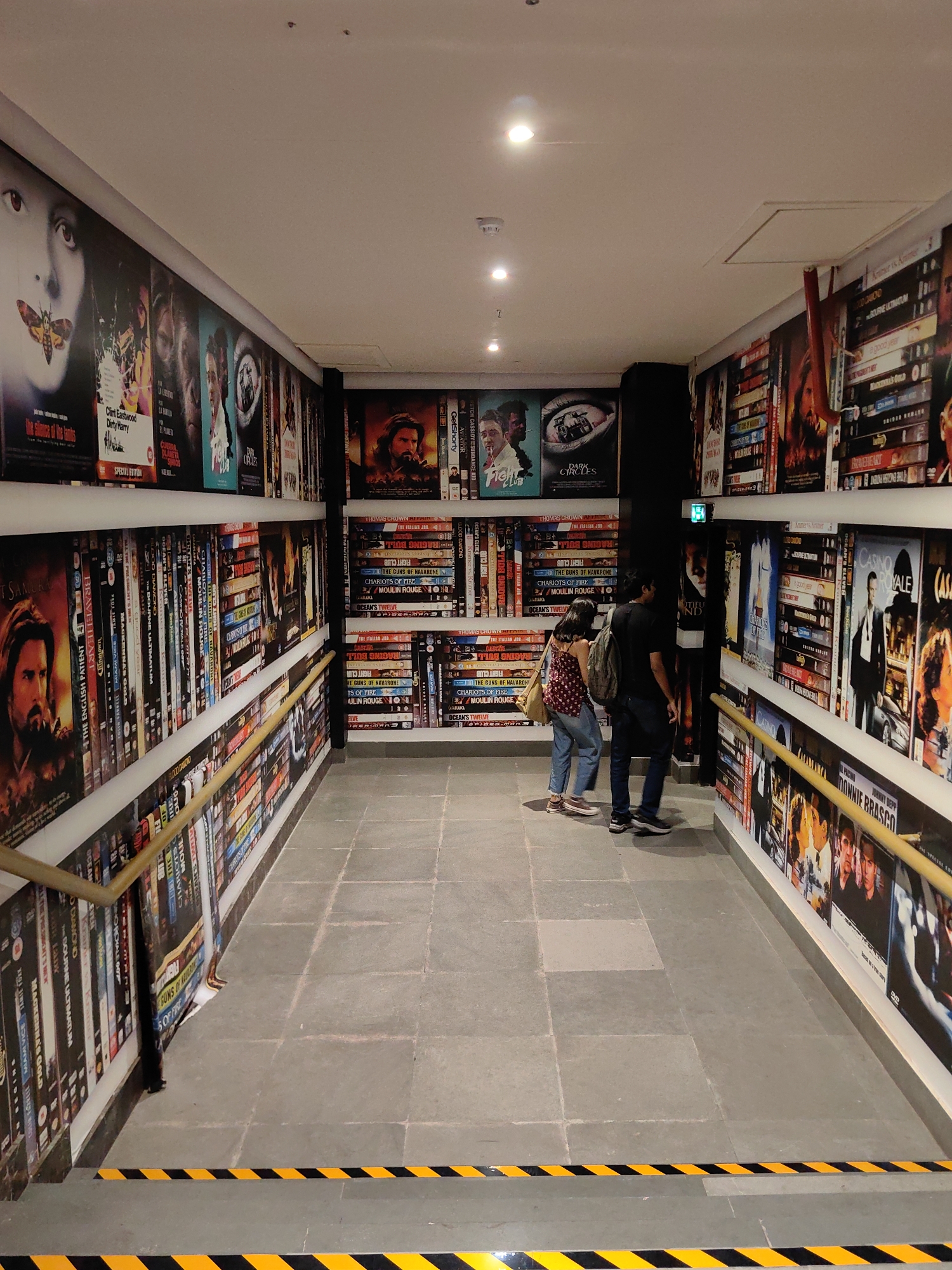 The image depicts an interior hallway lined with movie posters and DVD cases. Two individuals, a man and a woman, are walking down the hallway. The man is wearing a black t-shirt and dark pants, carrying a backpack. The woman is wearing a patterned top and jeans. The hallway walls are covered with numerous movie posters, including images related to "The Silence of the Lambs," "Clint Eastwood Dirty Harry," and "Casino Royale." Shelves on both sides of the hallway are filled with DVD cases. The floor is tiled with large gray tiles, and yellow and black hazard tape is visible on the floor at the bottom of the image. The lighting appears to be artificial, with several spotlights visible on the ceiling. This scene is located in the city of Mumbai, India, specifically at the Regal Cinema.