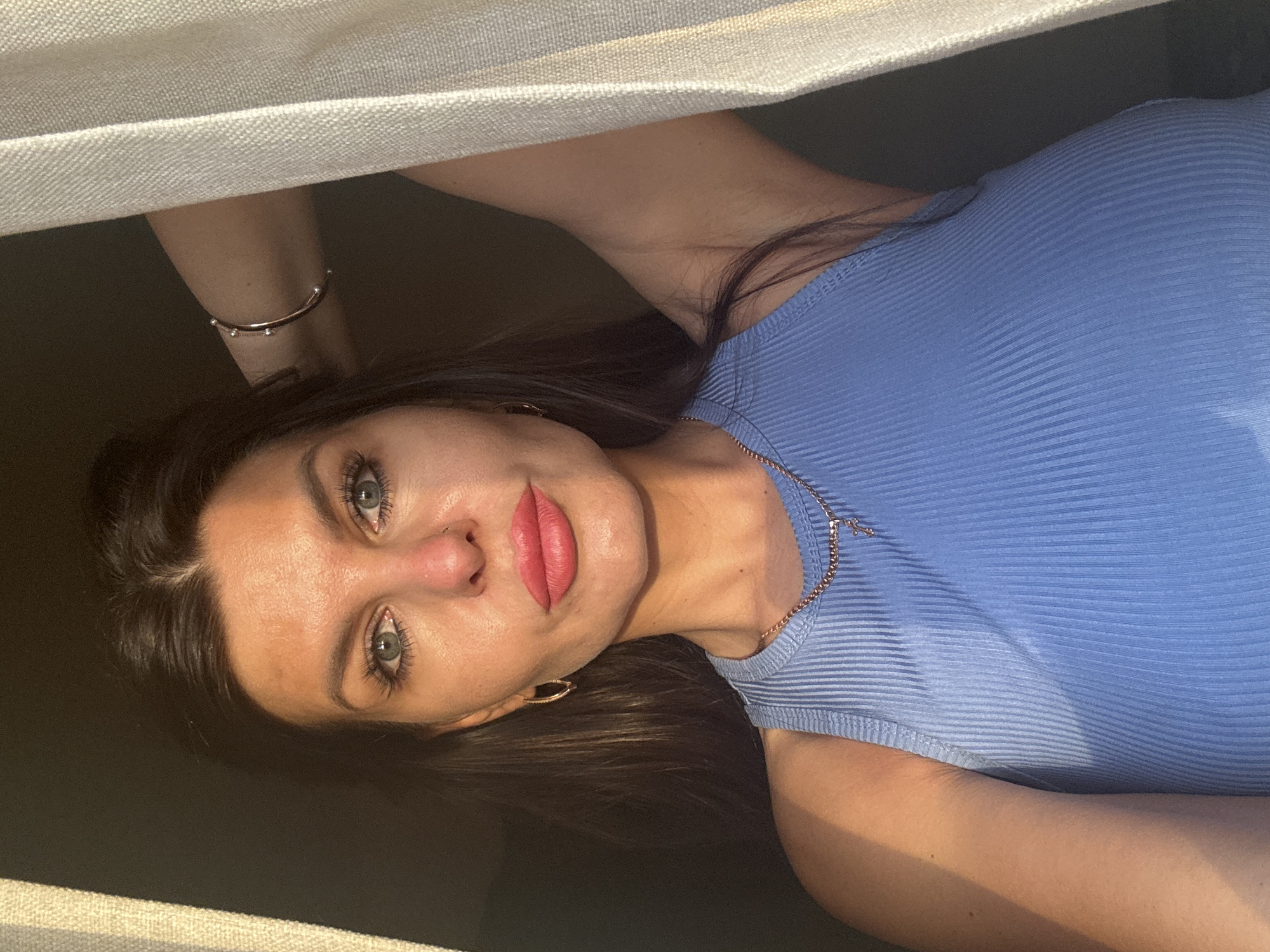 A close-up, medium-shot photograph captures a young woman from the chest up, positioned in front of a window with sheer curtains. The woman has fair skin, brown hair, and blue eyes. She is wearing a blue ribbed tank top and a rose gold necklace with a cross pendant. Her right arm is raised, resting on her head, revealing a rose gold bracelet on her wrist. The lighting suggests it is either sunrise or sunset, casting a warm glow on the left side of her face and body. The background is dark, with the curtains on the right side of the frame illuminated by the soft light. There is no identifiable location in the city of Chaikovsky, Russia, visible in the image.