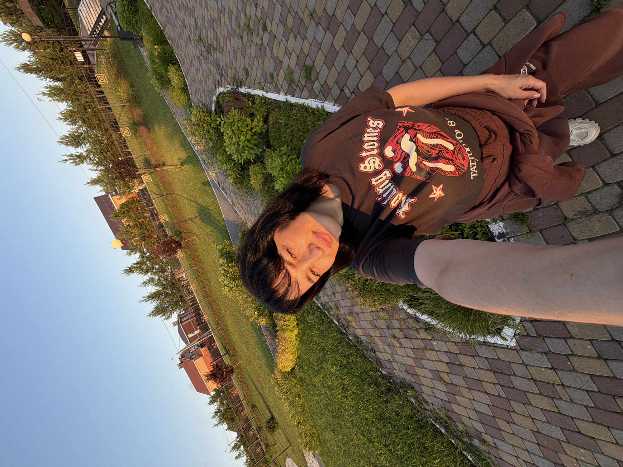 This media file is a selfie taken by a person outdoors, likely in a park or residential area in Shmidtovo, Russia. The image is oriented sideways.
The subject is a person with dark hair, wearing a dark t-shirt with a graphic print and brown sweatpants. They are looking at the camera, with a slight smile. Their right arm is extended, holding the camera. The visible text on the t-shirt reads "Rolling Stones Tattoo You".
The setting appears to be a paved walkway with diamond-shaped pavers, bordered by green grass and landscaping. In the background, there are trees, some residential buildings with red roofs, and a clear blue sky. The lighting suggests it is late afternoon or early evening, with the sun casting a warm glow. The overall mood of the image is casual and relaxed.
