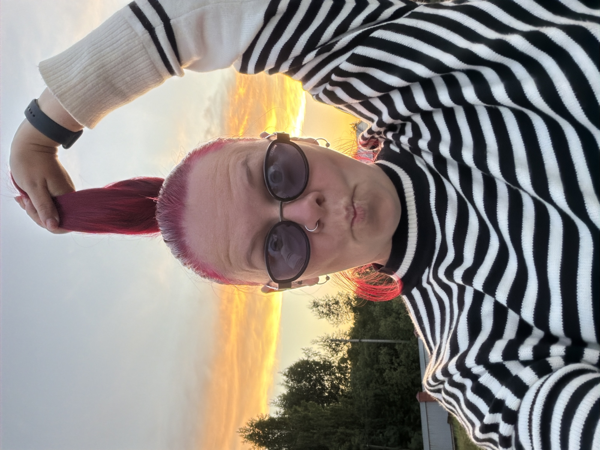 This image is a selfie of a person taken outdoors during sunset in Chaikovsky, Russia. The individual, seen from a low angle, has vibrant red or pink hair styled into a ponytail, which is held by their left hand. They are wearing a black and white horizontally striped knitted sweater and large, dark sunglasses. A septum piercing (nose ring) and earrings are visible on their face. A black smartwatch or fitness tracker adorns their left wrist.
The person has a casual expression with slightly puckered lips, typical of a selfie pose. The background features a dramatic sunset sky, dominated by warm orange and yellow hues near the horizon, transitioning to a lighter grey-blue overhead. Dark green trees are silhouetted against the bright sky along the horizon, suggesting a natural, possibly park-like or suburban, outdoor setting. The time of day is clearly late evening or dusk, highlighted by the glowing sky. The word "CELINE" is faintly visible on the arm of the sunglasses.