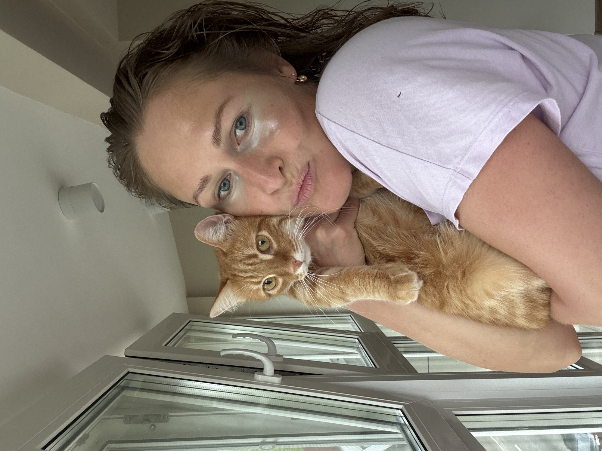 A woman with light skin, wet hair, and blue eyes is holding an orange tabby cat near a window. The woman wears a light purple t-shirt and gold earrings. The cat's fur is long and fluffy. The window is multi-paned with white frames and handles. The location is Chaikovsky, Russia, based on external data not available in the image. A white cylindrical object is visible on the wall near the top left corner. The woman and cat are close together, suggesting affection. The angle of the photo is slightly tilted, looking down on the scene.