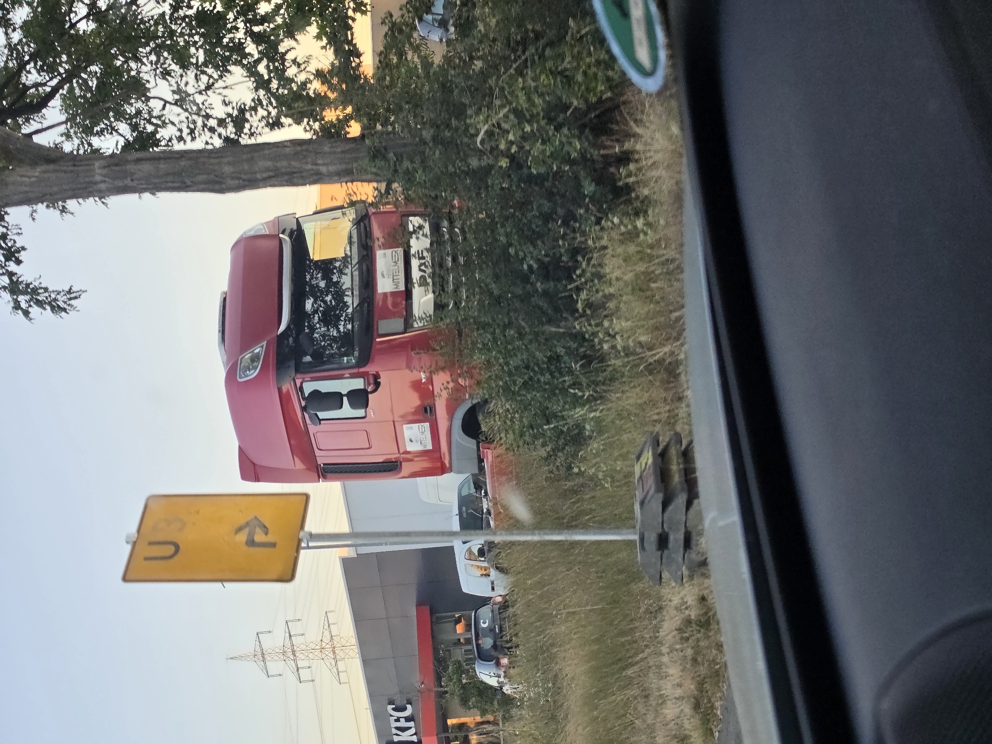 The image shows a red DAF semi-truck parked on a grassy area near a KFC restaurant. A yellow directional sign with "U3" and a right arrow is visible to the left of the truck. In the background, a large electrical pylon and a building with a KFC sign are present. Several other vehicles, including a white van and a dark-colored car, are parked in front of the KFC. The scene appears to be a roadside area, possibly a rest stop or a parking lot adjacent to the restaurant. The overall lighting suggests it is either early morning or late afternoon.