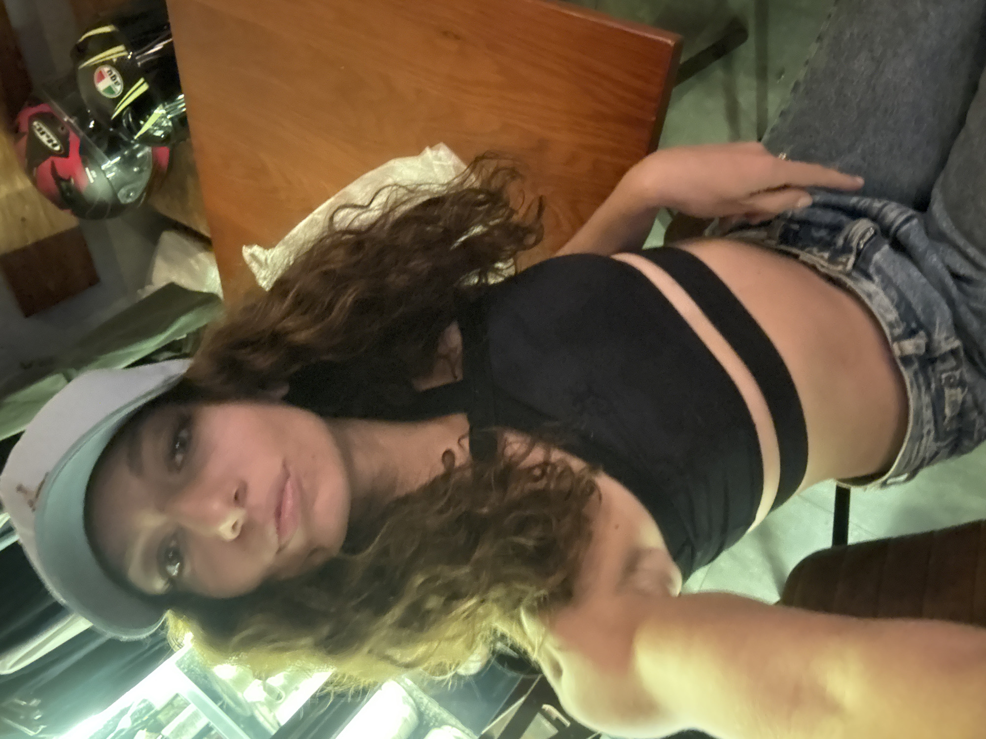 A woman is lying on her back, angled with her head towards the left and her legs towards the right of the frame. She is wearing a light blue baseball cap, a black crop top with straps, and light blue denim jeans. The visible skin on her midriff and between her top and jeans suggests that she is wearing the jeans low. She has long, curly brown hair. Her gaze is directed toward the camera. A wooden table is visible above her head, with two motorcycle helmets near the upper left corner of the frame. The background is a mixture of blurred objects, including chairs and the floor. The location is not specified, and information about Đà Nẵng, Vietnam is not evident in the image.