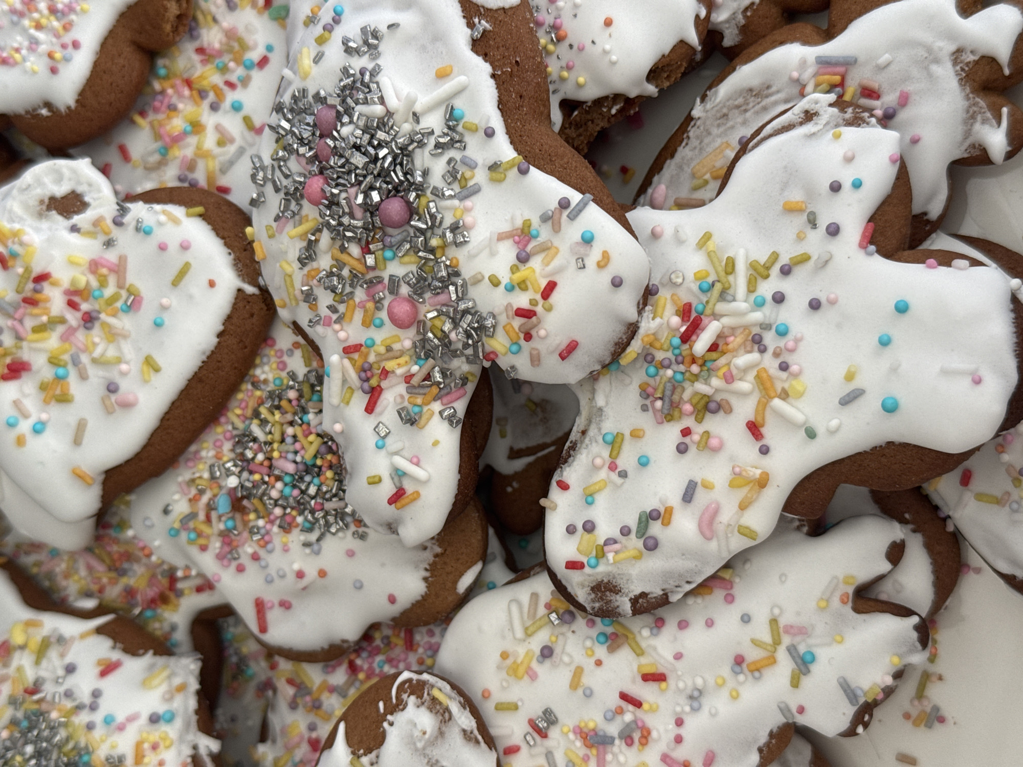 The image shows a close-up of several gingerbread cookies. The cookies are decorated with white icing and a variety of colorful sprinkles. Some sprinkles are elongated, while others are shaped like small spheres. There are also silver sprinkles. The cookies are piled on top of each other. The image doesn't provide any specific information about the location or the city of Šušanj, Montenegro.