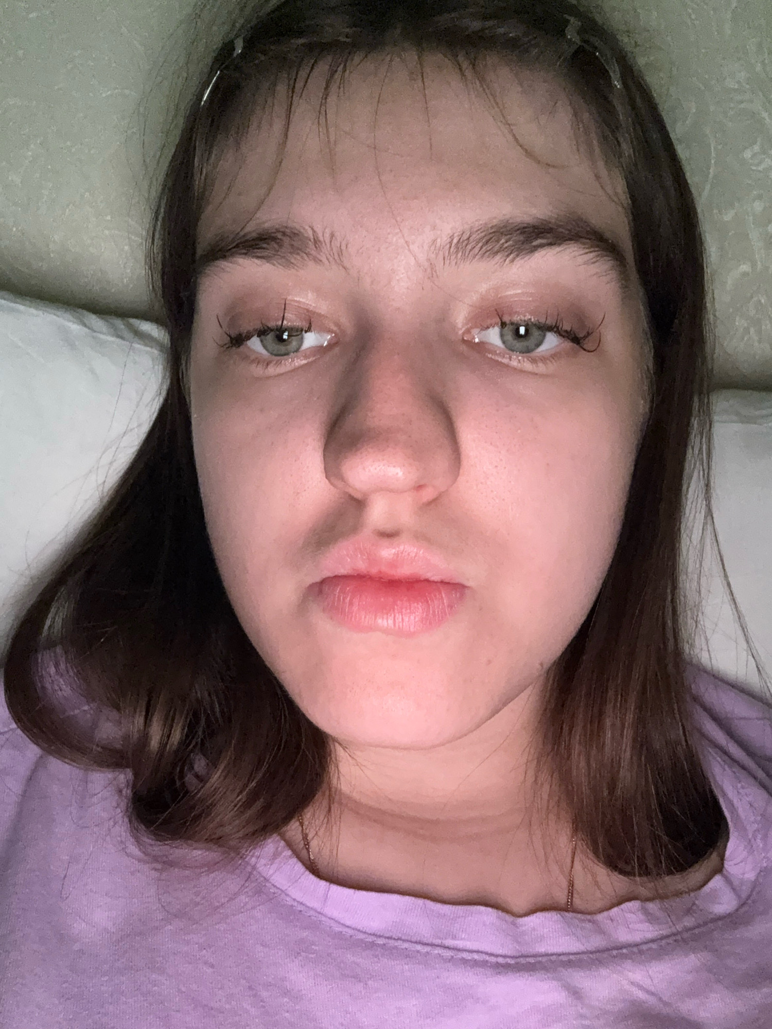 This is a headshot of a young woman with brown hair and light green eyes, wearing a purple t-shirt. She is lying down, possibly in bed, as a pillow is visible behind her head. The background appears to be a textured wall. The lighting is dim, suggesting an indoor setting. There are no discernible landmarks or environmental cues to indicate a specific location within Zavolzhye, Russia.
