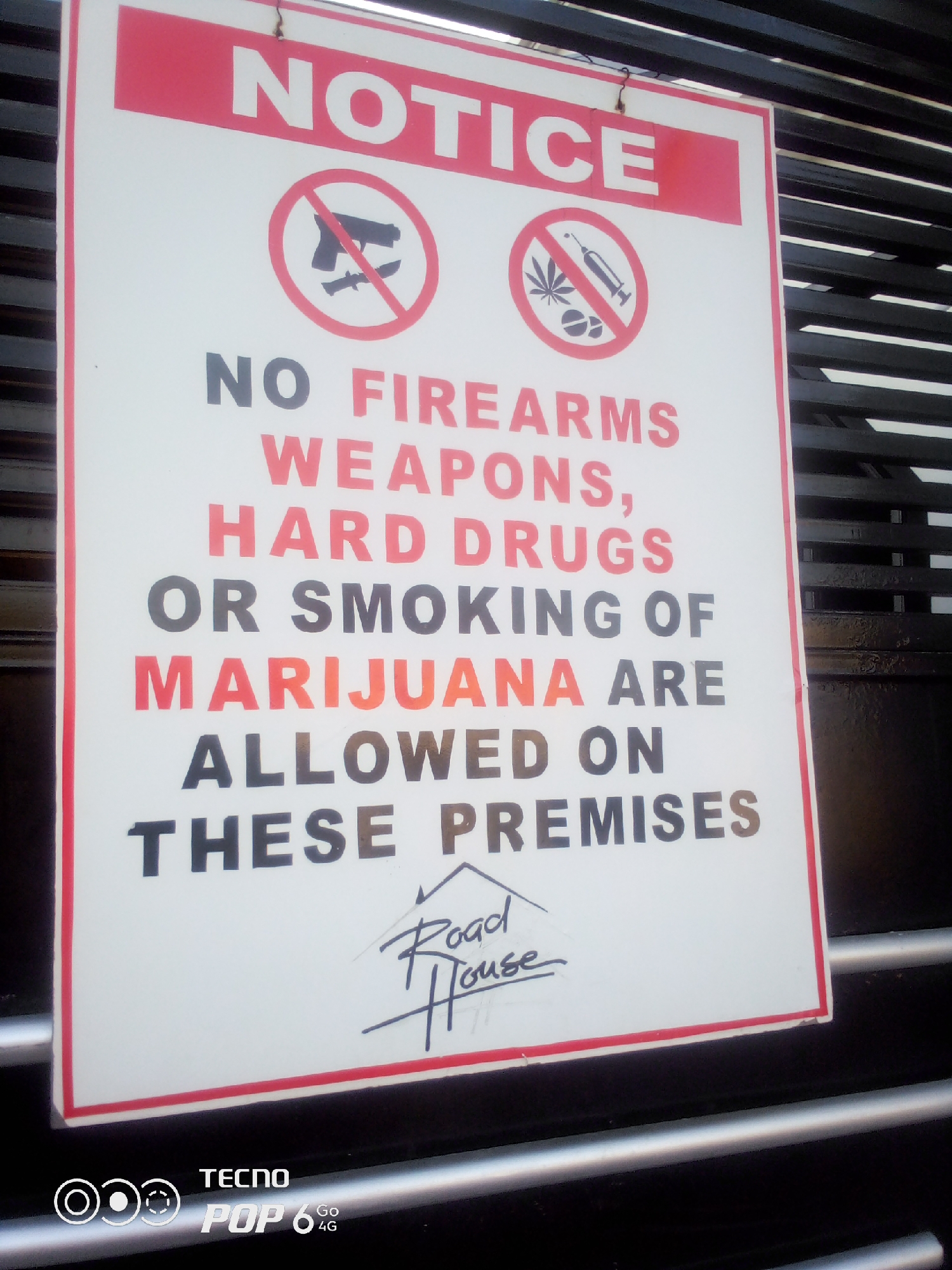 The image shows a "NOTICE" sign, prominently displayed outdoors, likely at the entrance of a establishment. The sign is white with a red border and red text at the top. It features two circular icons with a red border and a red diagonal line, indicating prohibition. The left icon depicts a gun and a knife, while the right icon shows a marijuana leaf and a syringe. Below these icons, the sign states in large letters: "NO FIREARMS, WEAPONS, HARD DRUGS OR SMOKING OF MARIJUANA ARE ALLOWED ON THESE PREMISES". At the bottom of the sign, there is a stylized handwritten signature that appears to read "Road House". The sign is mounted against a dark, slatted background, possibly a fence or gate. The bottom of the image shows a watermark for "TECNO POP 6 4G". There are no people or other visible subjects in the image. The overall setting is an exterior urban environment, likely in the city of Howayi, Nigeria, given the context.