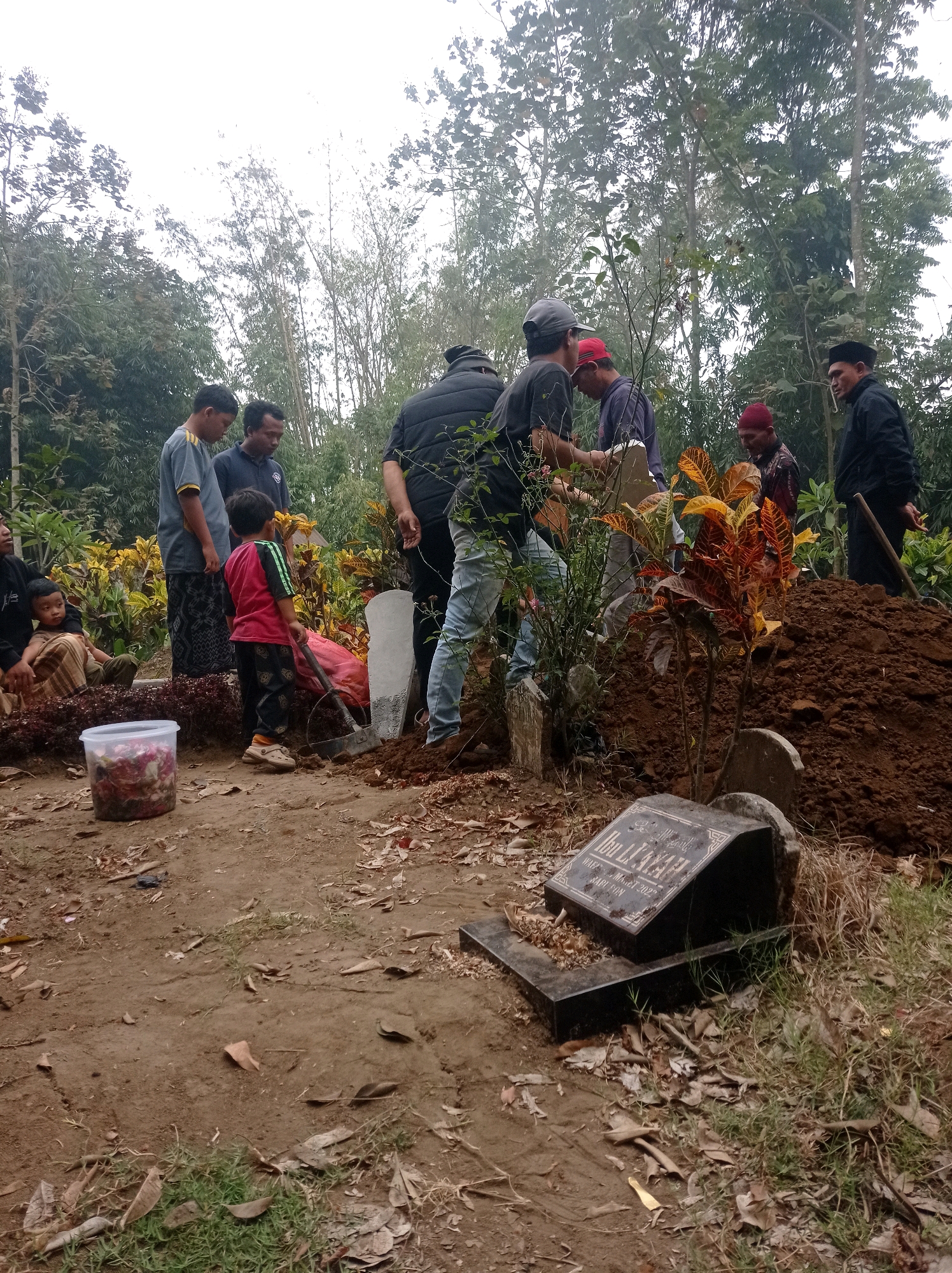 The image depicts a group of people gathered around a freshly dug grave in a cemetery located in Pandanmulyo, Indonesia. The scene appears to be a funeral or burial service. Several individuals are dressed in dark clothing, common for such occasions, while others are in casual attire. A young child in a red shirt and black pants stands near the grave, and another child sits on the ground to the left.
In the foreground, a black headstone is partially visible, bearing Arabic script and the date "March 2022." A pile of dark soil indicates recent excavation. A red plastic bag and a shovel lie near the grave, suggesting ongoing or recently completed burial activities. A clear plastic container filled with what appears to be flower petals is also present.
The environment is outdoors, surrounded by trees and dense foliage, characteristic of a rural or semi-rural setting. The ground is a mix of dry earth, fallen leaves, and sparse vegetation. The sky is overcast, contributing to a somber atmosphere. The overall scene suggests a communal act of remembrance and farewell.