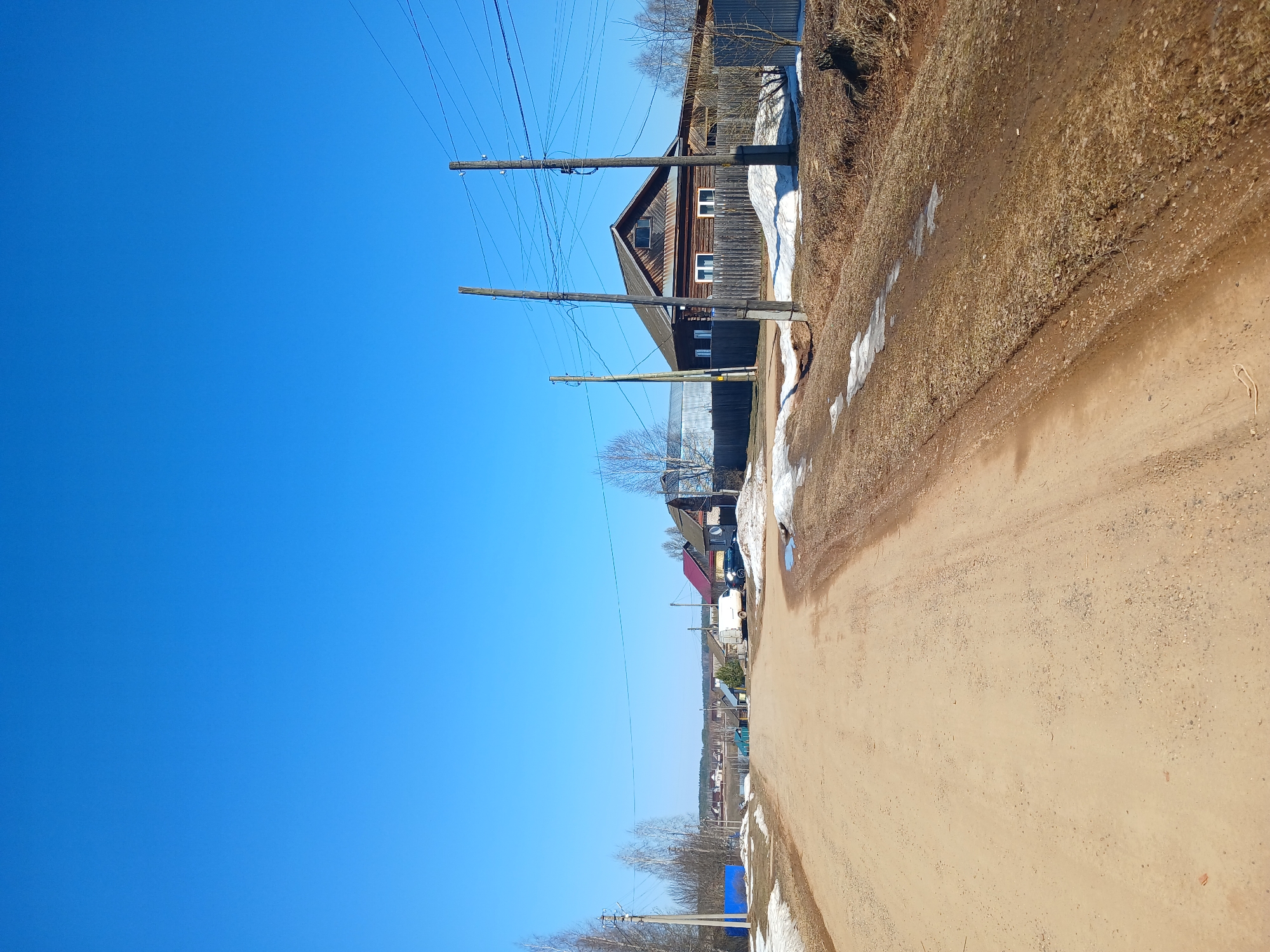This image depicts a tranquil rural village scene, likely during late winter or early spring due to the presence of melting snow and bare trees. A wide, unpaved dirt road dominates the right side of the frame, extending from the foreground into the distance. Along the left side of the road, several wooden houses and buildings are visible.
The most prominent structure is a two-story wooden house with a dark brown facade, white-framed windows, and a distinctive gabled roof, situated in the mid-ground. Parallel to the road and the houses, several wooden utility poles stand tall, supporting numerous overhead electrical wires that crisscross against the sky. The ground bordering the road is covered with dry, brownish grass and weeds, punctuated by lingering patches of white snow.
In the mid-distance, a white vehicle, possibly a van, and another dark vehicle are parked near one of the houses. Further down the road, a blue tarp or covering is visible on the ground. The sky above is a vast, clear, and uniformly bright blue, indicating sunny weather.
No people are visible, and no specific activity is taking place, creating a static and peaceful ambiance. The bright light suggests it is midday or early afternoon. There is no visible text. The overall scene conveys a sense of quiet rural life in Bolshaya Sosnova, Russia, as winter transitions to spring.