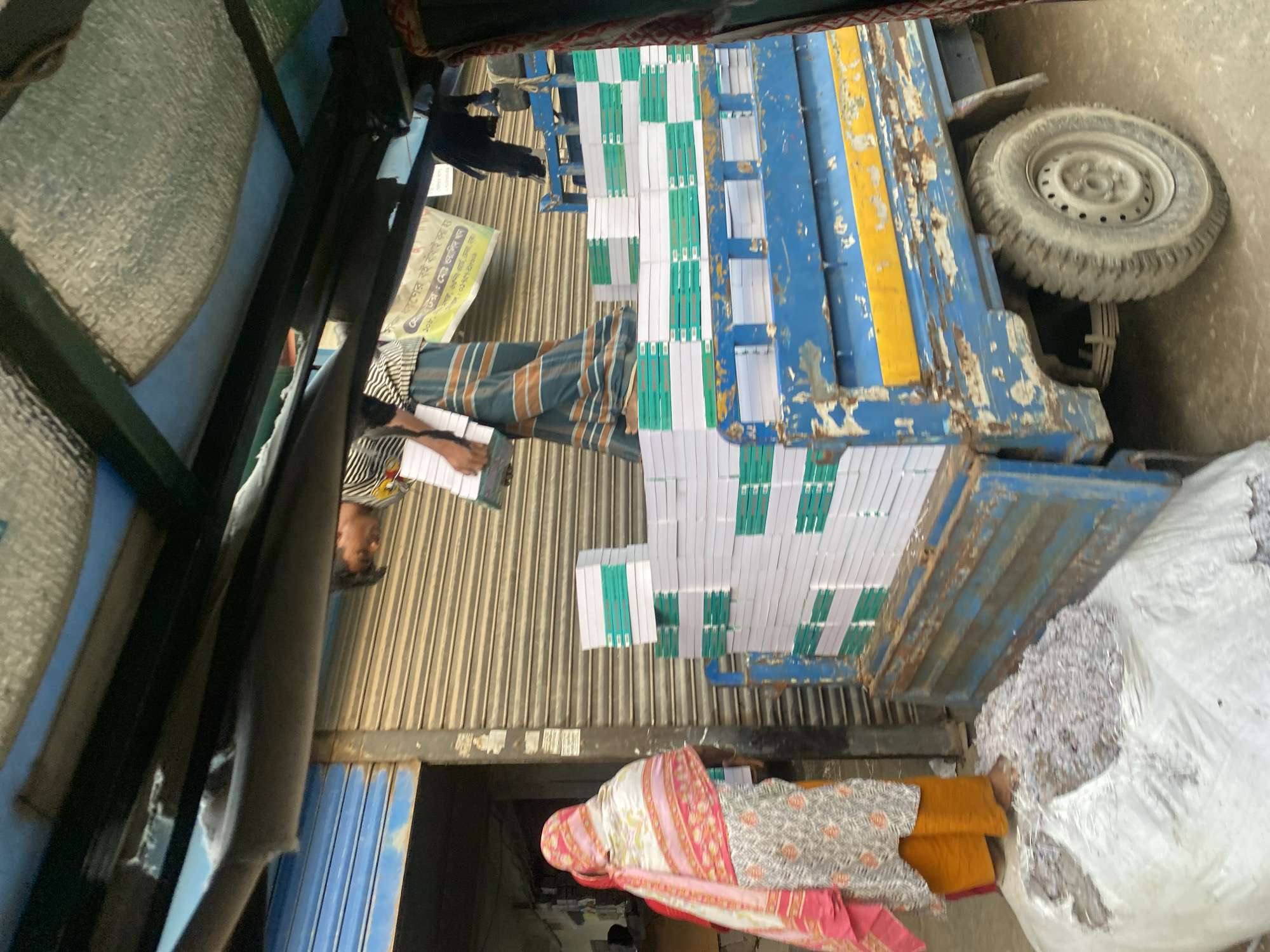 The image shows a street scene in Dhaka, Bangladesh. The foreground is dominated by a blue delivery truck loaded with stacks of notebooks wrapped in white paper with green trim. A large white sack filled with shredded paper sits beside the truck.
A man in a striped shirt and lungi is standing by the truck writing on some papers. A woman in a shawl and yellow trousers stands nearby. A poster with Bengali script is partially visible near the man, mentioning "Walid Khan" and a phone number, suggesting a business related to bookbinding. The background shows corrugated metal and other shop fronts typical of a busy urban street.