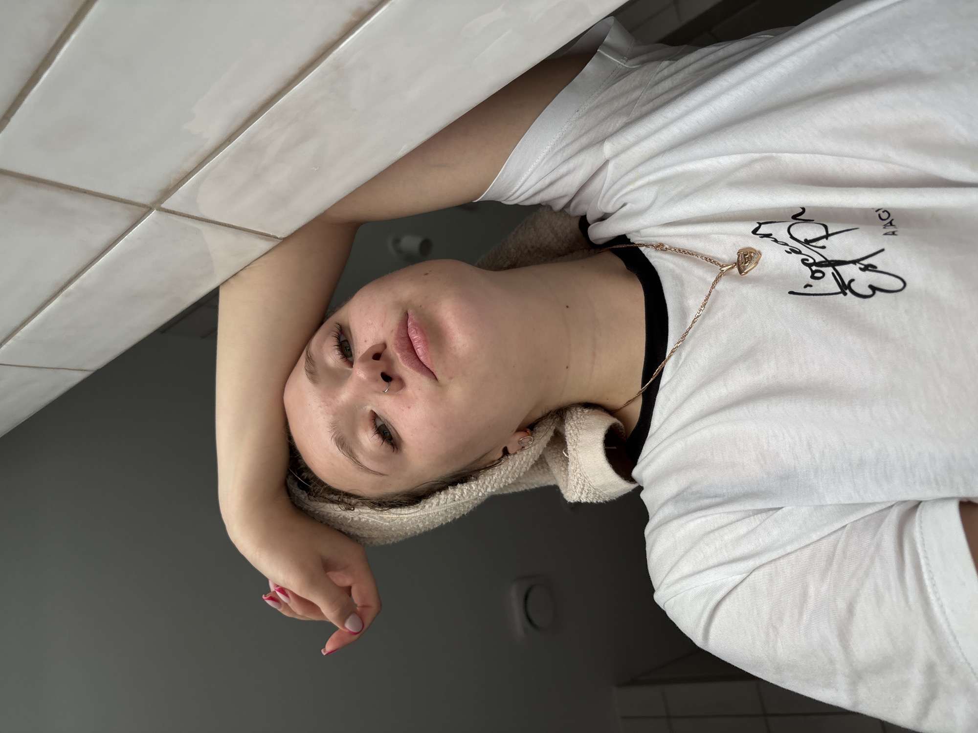 A young person with light skin is leaning their head against a white, tiled wall. They have a beige towel wrapped around their head. They are wearing a white T-shirt with black trim and a gold necklace with a heart-shaped pendant. The background is a gray-toned wall and floor. It is not possible to determine the location as Izhevsk, Russia from the given image.