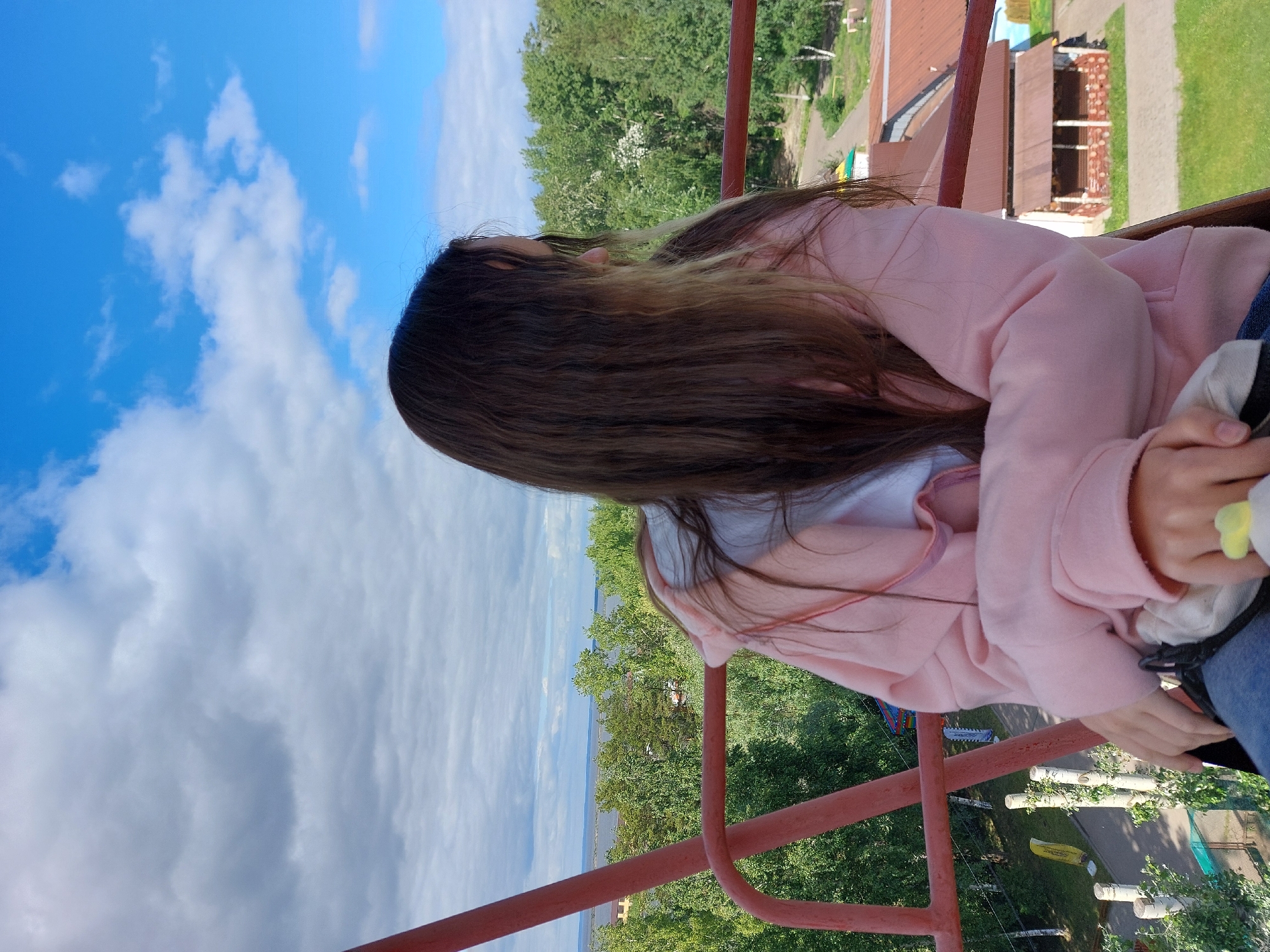 A young woman with long, brown hair, partially illuminated by sunlight, is seated in what appears to be a Ferris wheel carriage, looking out to her right. She is wearing a pink hooded jacket and blue jeans. Her hands are resting in her lap. The carriage has a red metal frame. In the background, a landscape is visible, featuring a body of water, lush green trees, and some buildings with brown roofs. The sky above is a vibrant blue with scattered white clouds. This vantage point suggests the Ferris wheel is located in an area with a view of natural scenery and some urban or semi-urban development, consistent with a park or amusement area within Chaikovsky, Russia, possibly overlooking the Kama River.