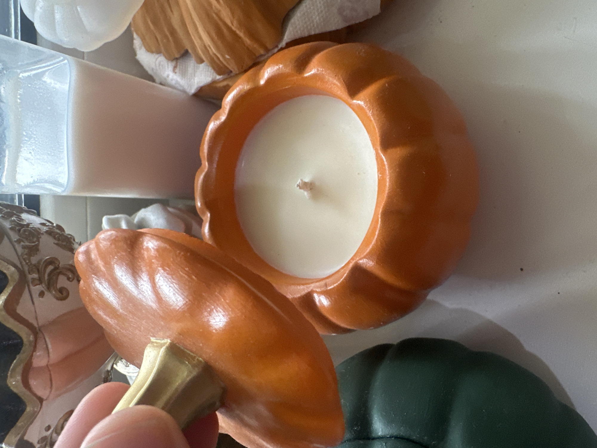 An orange pumpkin-shaped candle is in the foreground, with its lid being held by a finger and thumb. The candle itself is white wax with a wick. To the left, a dark green pumpkin-shaped object is partially visible. In the background, a clear container with a light-colored liquid, possibly milk or lotion, is present, along with other decorative pumpkins. The surface appears to be a countertop or table. No location in Vladivostok, Russia can be determined from the image.