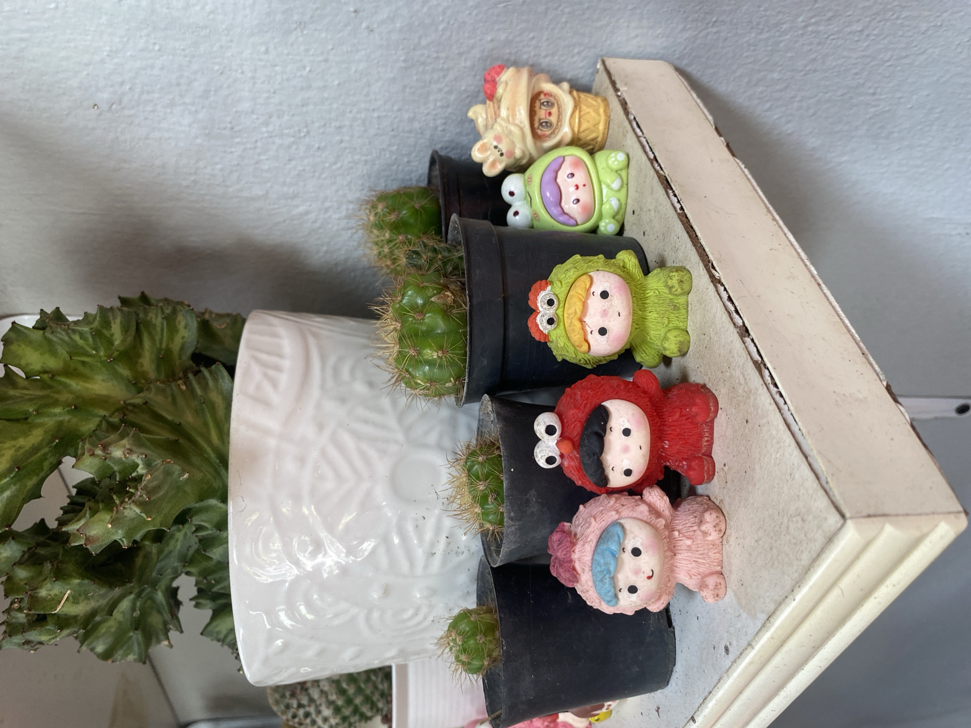 A still life photograph shows several small cacti in black plastic pots arranged on a shelf. On top of each pot is a small, cartoonish figurine. From top to bottom, the figurines are shaped like an ice cream cone with a bunny, a purple and green alien-like creature, a green monster, a red Elmo-like character, and a pink bear-like character with a blue bow. To the left, a larger white decorative pot contains a green leafy plant. The shelf is white and shows signs of wear. The photograph was taken indoors. This photograph may have been taken in Choeng Thale, Thailand, but there is no specific information to confirm this.