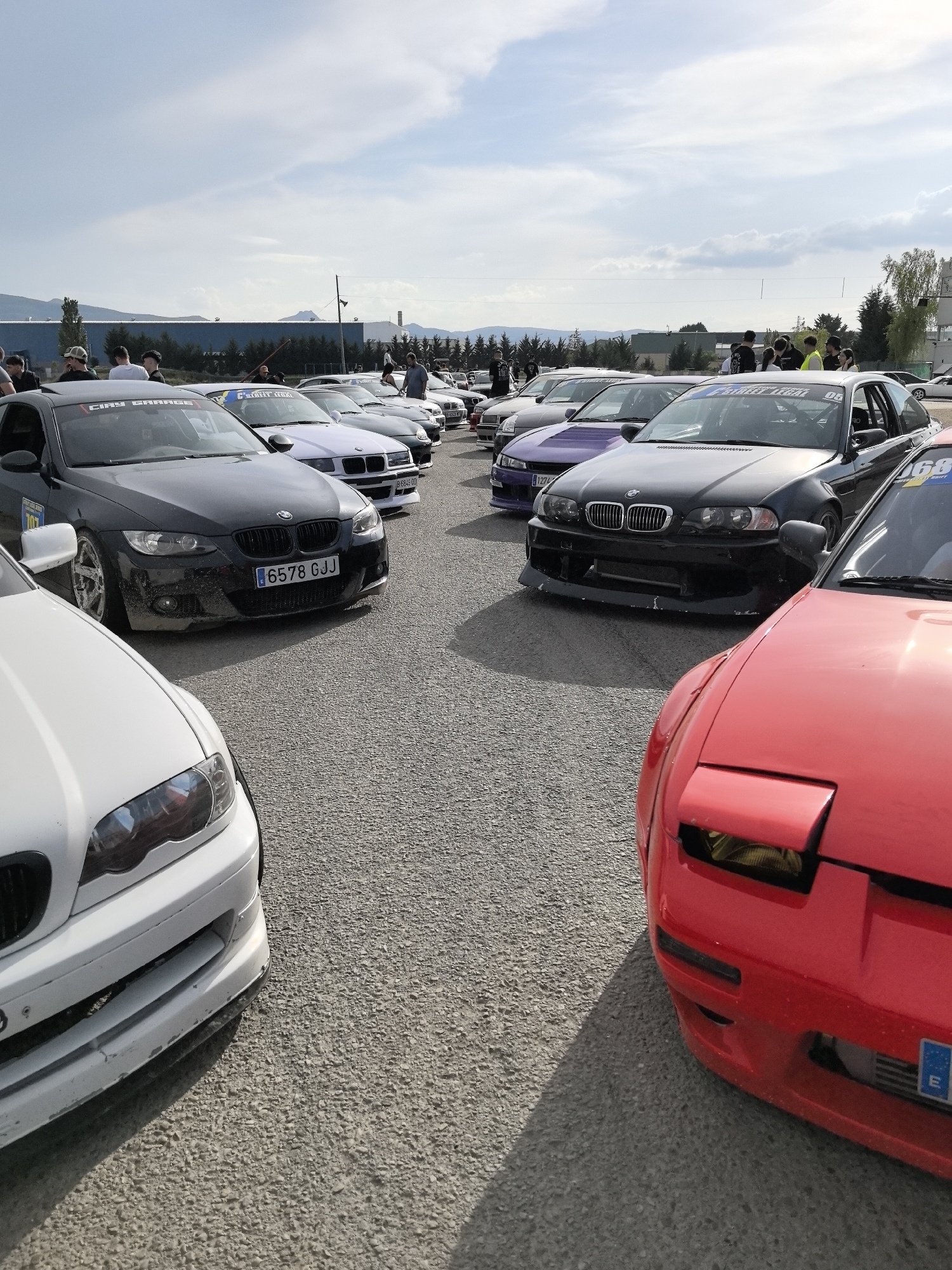 The image shows a gathering of modified cars in what appears to be a parking area. Several BMWs of the E46 and E90 generations are present, alongside a red Nissan 240SX (S13). A purple Nissan Silvia S14 can also be seen. Most of the cars have modifications suggesting a drift or track racing use, such as aftermarket bumpers, lowered suspension, and possibly roll cages. People are standing around and between the cars, likely car enthusiasts. The license plate "6578 GJJ" is visible on one of the BMWs. The location appears to be on the outskirts of a town or city, given the presence of industrial buildings and distant mountains in the background. Based on the license plate formats and overall environment, the image appears to be taken in Spain. The presence of mountains and industrial buildings suggests the location is likely in or near Miranda de Ebro, Spain.