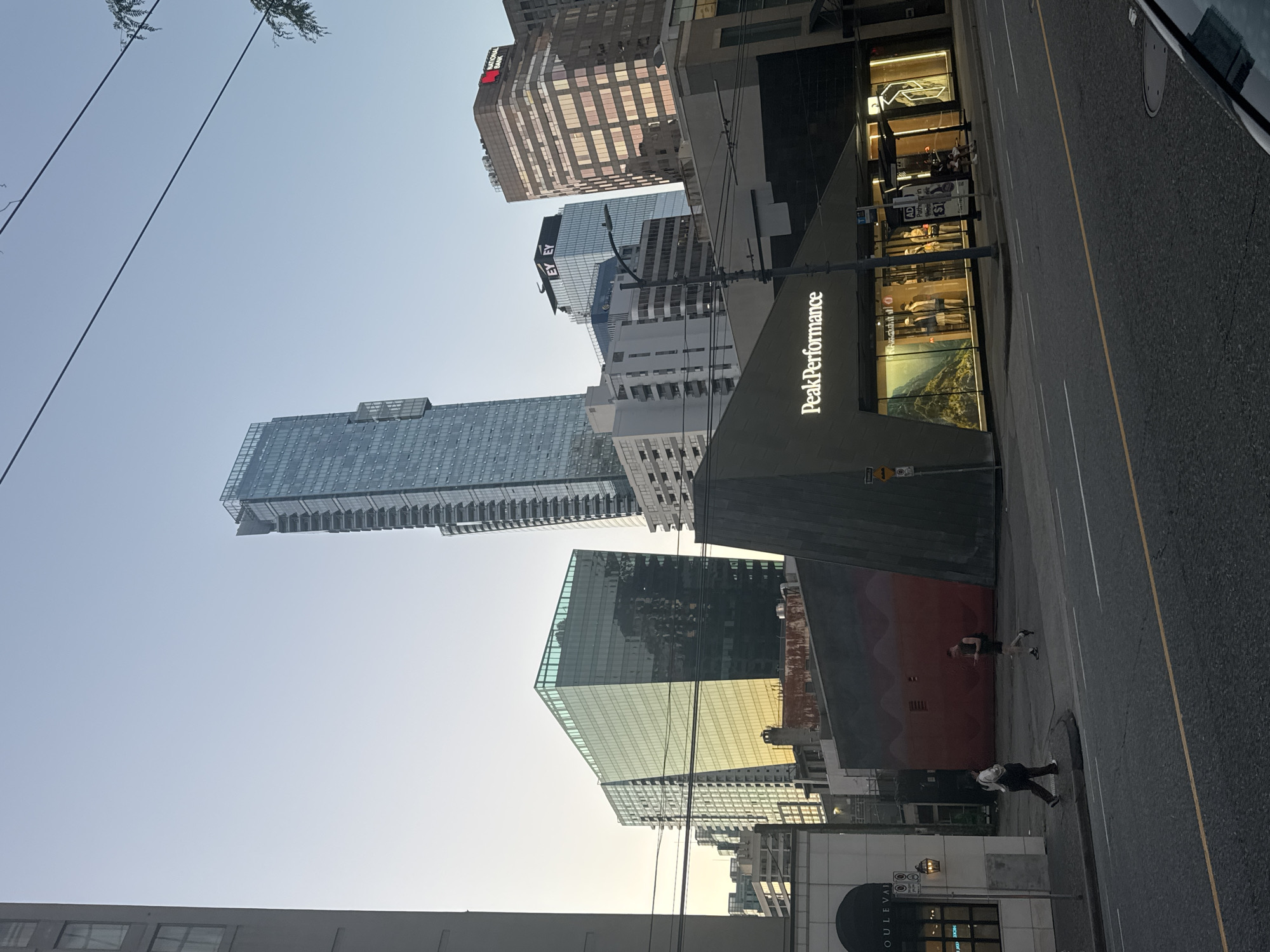 The image depicts a street scene in Vancouver, Canada, with a mix of high-rise buildings dominating the skyline. The prominent Peak Performance store is visible on the right side of the street. Two individuals are walking on the sidewalk, one appears to be running. The environment includes a street with lane markings, sidewalks, and visible street furniture. Buildings of varying heights and designs line the street, with some showing reflective glass facades. Overhead wires are also visible.