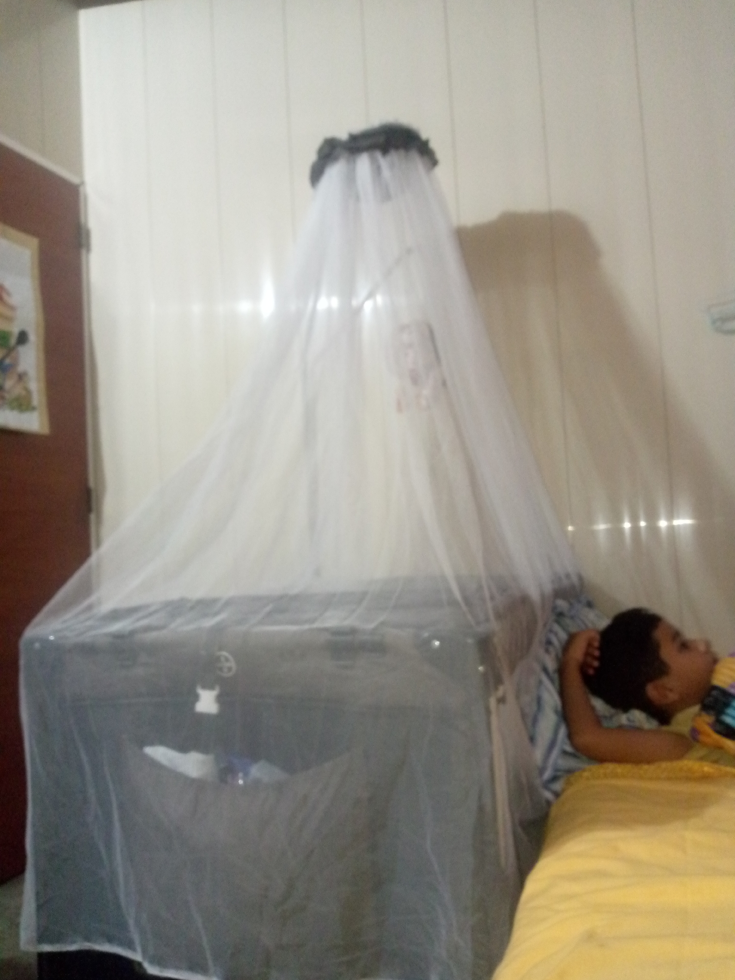 An indoor scene in Altagracia, Venezuela, features a young child resting on a bed next to a large crib or playpen covered by a white mosquito net.
On the left side of the frame, a brown wooden door is partially visible, next to which hangs a framed picture on the light-colored wall. The walls throughout the room are paneled with vertical strips.
Dominating the center is a substantial baby crib or playpen, completely enclosed by a translucent white mosquito net. The net is draped from a circular top structure, forming a canopy over the crib. Inside the crib, indistinct items and a white, soft object are visible through the netting. A small white figure, possibly a teddy bear or toy, is attached to the exterior side of the crib.
To the right of the crib, a child, appearing to be a young boy, is lying on a bed. His head is propped up by his right arm, and he looks calmly towards the crib. He wears a striped shirt with yellow and other colors. The bed is covered with a soft, yellow sheet or blanket.
The room is illuminated by what appears to be natural light, with bright reflections visible on the paneled wall behind the crib and child, suggesting it is daytime. The presence of a mosquito net indicates a practical measure against insects, common in tropical climates. No visible text is present in the image.