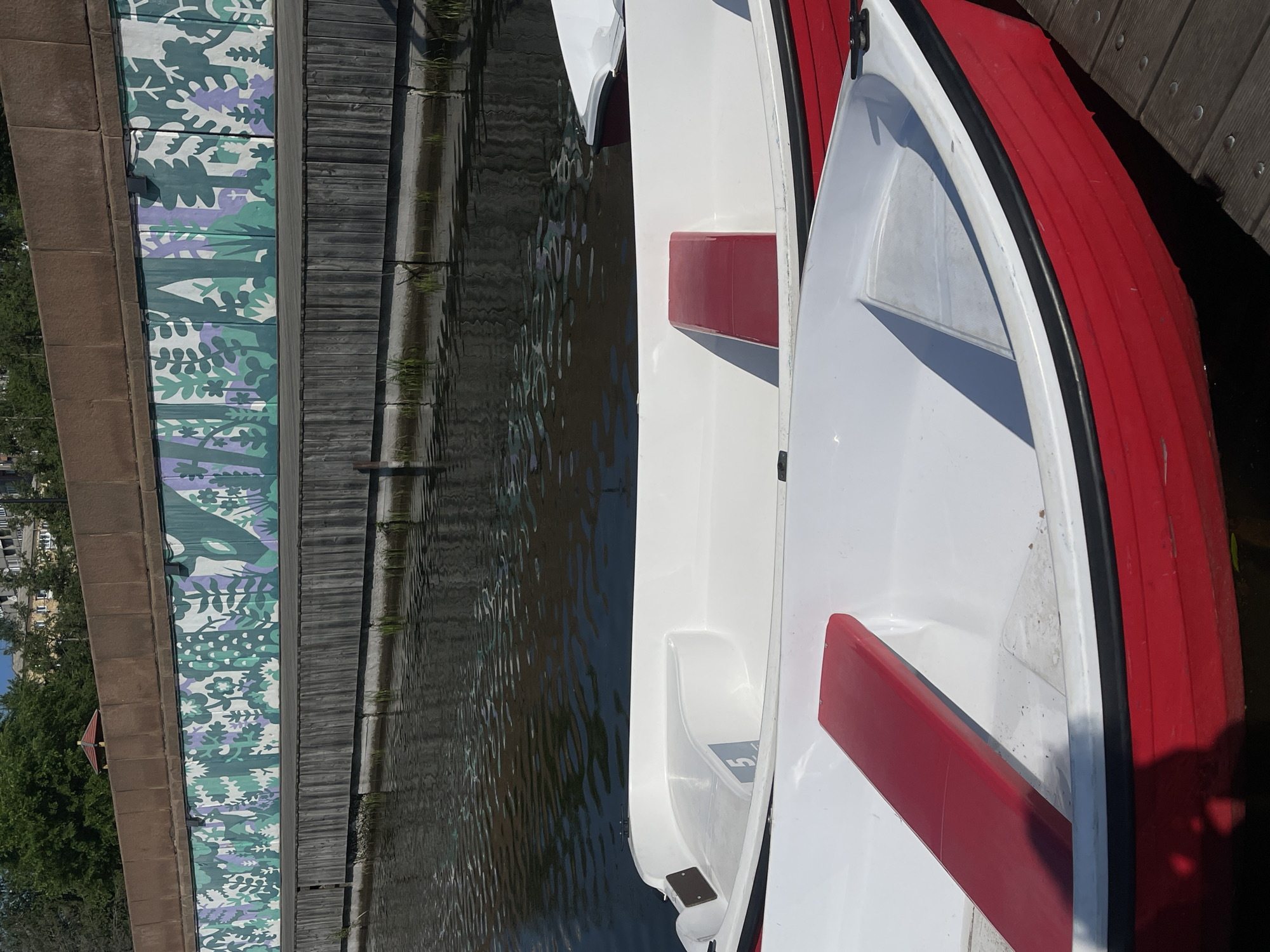 The image captures a serene waterside scene. In the foreground, two white boats with red accents are docked on a wooden pier. The boat in the lower half of the frame is predominantly red with a white interior, while the boat partially visible above it is mostly white with a red seat. The water is calm and reflects the surrounding environment. Behind the boats, a wooden fence runs along the water's edge, topped by a wall adorned with a colorful mural depicting stylized plants and foliage in shades of green, purple, and white. In the background, trees and buildings can be seen, suggesting an urban setting. This scene is located at the embankment of the Yenisei River in Krasnoyarsk, Russia, specifically the embankment near the Staryi Dom restaurant, a popular recreational spot.