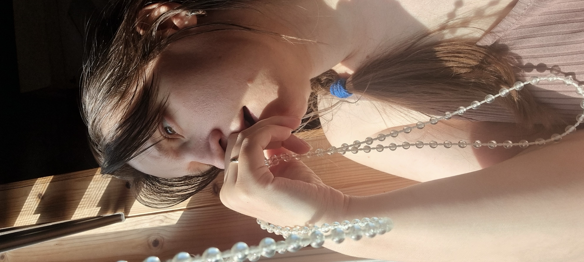 The image presents a close-up, angled view of a person indoors, featuring their face, hand, and hair. The person has long, dark brown hair, which is tied back with a blue elastic band in a ponytail. Their fair-skinned face is partially visible, showing one brown eye with a direct, intense gaze, and part of their nose and mouth area.
The person is holding a long strand of clear, spherical beads with their right hand, wearing a silver-toned ring on one finger. The beads appear to be a necklace or a rosary. The background consists of a light-colored wooden surface, possibly a floor or a table, bathed in bright, directional sunlight. This sunlight creates strong, distinct shadows, suggesting it is daytime, likely morning or late afternoon. A dark, slender object, possibly a pen or part of a window frame, is visible on the wooden surface in the lower left. The person is wearing a light-colored, possibly ribbed, garment. There is no visible text in the image. The overall impression is a contemplative moment captured in natural light. The setting is indoors, with the provided context indicating Dunnae-myeon, South Korea.