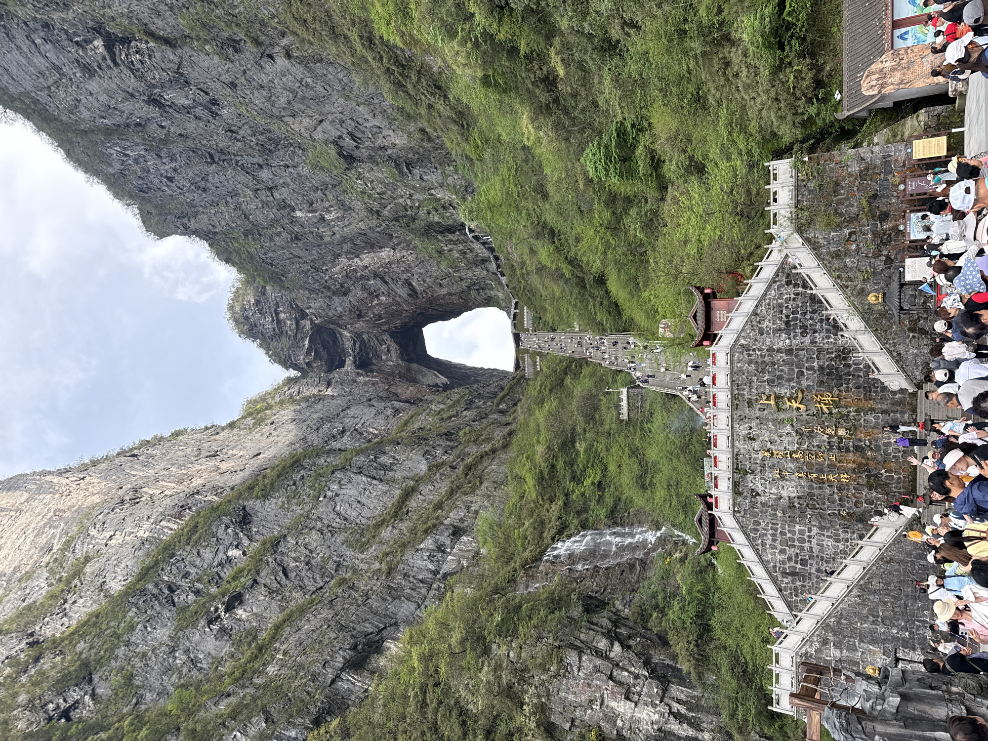 The image depicts Tianmen Mountain in Zhangjiajie, China, featuring a large natural archway. A crowd of people are gathered at the mountain's base, near a stone structure with Chinese writing. A staircase leads up the mountain. A narrow pathway with people traversing it extends to the archway. The landscape is dominated by steep, rocky cliffs covered with vegetation. A waterfall cascades down one of the cliffs. The sky above the archway is visible with clouds.