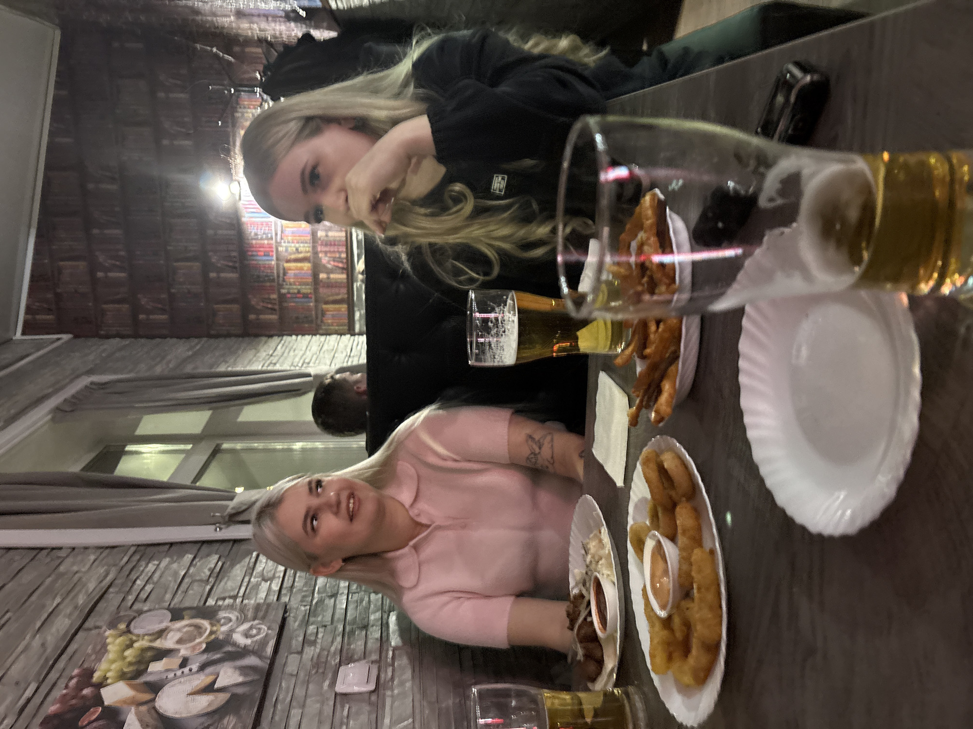 The image depicts two young women seated at a table, likely in a restaurant or bar. The woman on the left is wearing a pink sweater, while the woman on the right is wearing a black top. They appear to be in a social setting, possibly dining together.
On the table, there are several plates of food, including what appear to be fried foods (possibly french fries and onion rings) and a small dish with a dip or sauce. There are also glasses filled with beer or a similar beverage.
The background shows a brick wall, suggesting an interior setting. The presence of artwork on the wall and visible window frames add to the impression of a casual dining environment. The image is taken at a low angle, creating a somewhat distorted perspective of the table and its contents.
There is no verifiable information to conclude the location is Ust-Ilimsk, Russia.