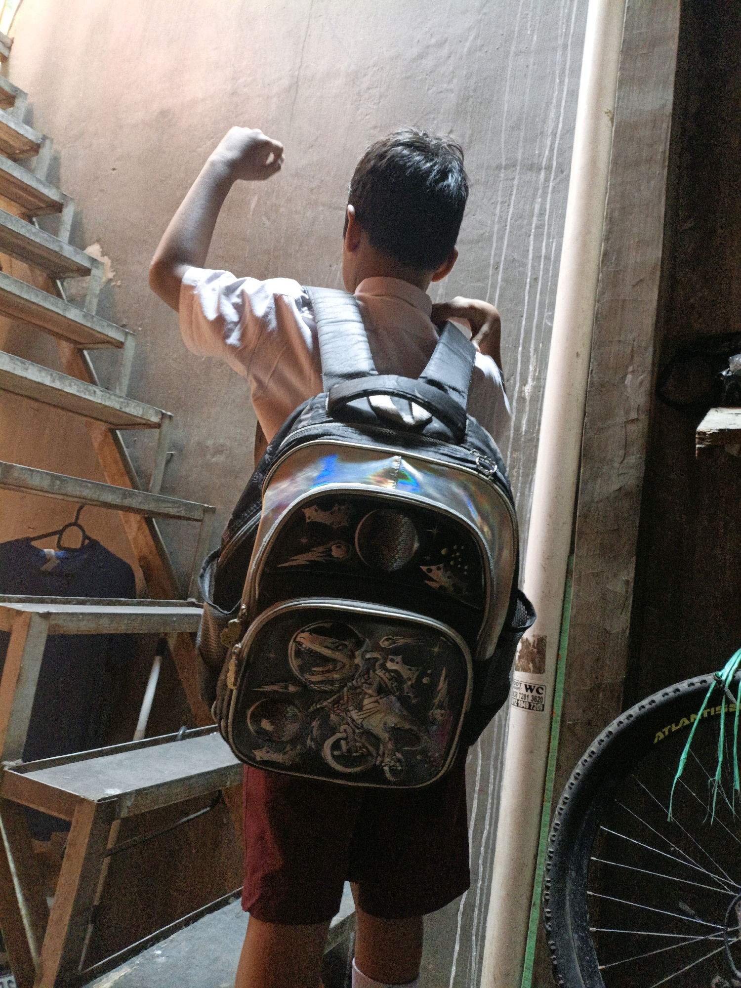 A boy wearing a white shirt and red shorts with a silver and black backpack is standing on a staircase. The boy is facing away from the camera and appears to be flexing his arm. The staircase is made of metal and wood and is located indoors. There is a white pipe running along the wall. A bicycle wheel is visible on the right side of the image. The scene appears to be in a residential area of West Jakarta, Indonesia, likely within a modest home or building, given the unfinished concrete walls and simple staircase construction.
