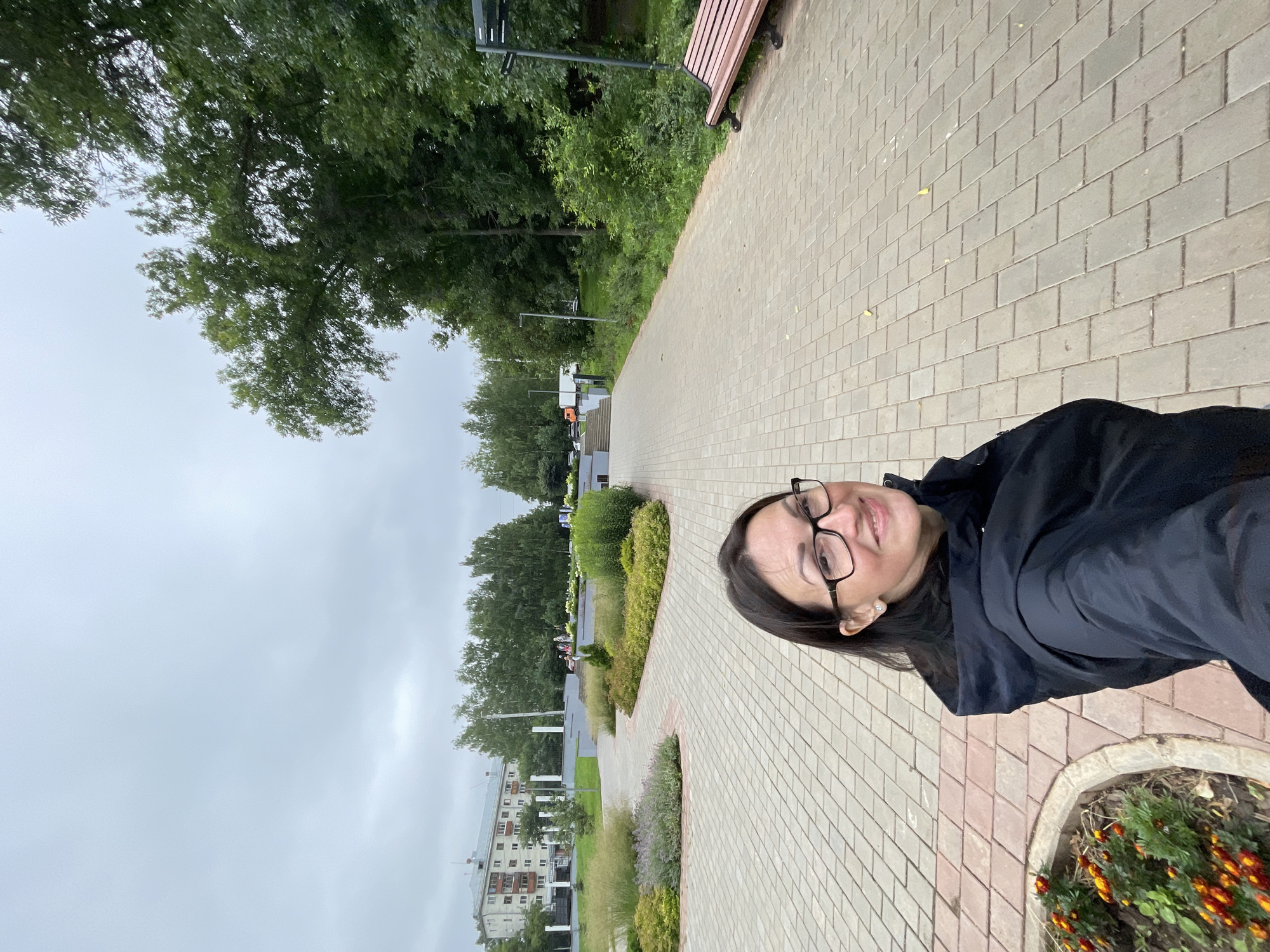 A selfie is taken by a woman with dark hair and glasses in a black jacket, standing on a paved walkway in a park in Kirov, Russia. The walkway is made of light brown rectangular pavers and curves to the right. To the left of the walkway, there is a flower bed with orange and yellow flowers. Further to the left, there is a landscaped area with green shrubs, purple flowers, and ornamental grass, and behind that, a multi-story apartment building. Tall green trees line the right side of the walkway, and a park bench is visible in the distance. The sky is overcast and gray.