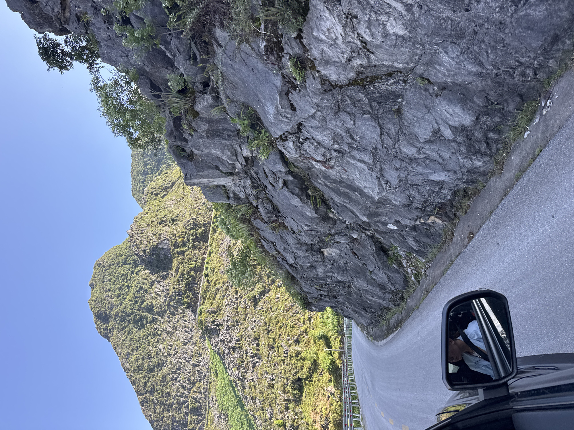 The image depicts a road winding through the mountainous landscape of Đồng Văn Commune, Vietnam. A large, gray, rocky cliff face dominates the right side of the frame, with vegetation growing on its surface. A guardrail runs along the edge of the road on the left, separating it from a steep, green hillside covered in trees and shrubs. The sky is clear blue. In the foreground, the side mirror of a vehicle reflects the driver.