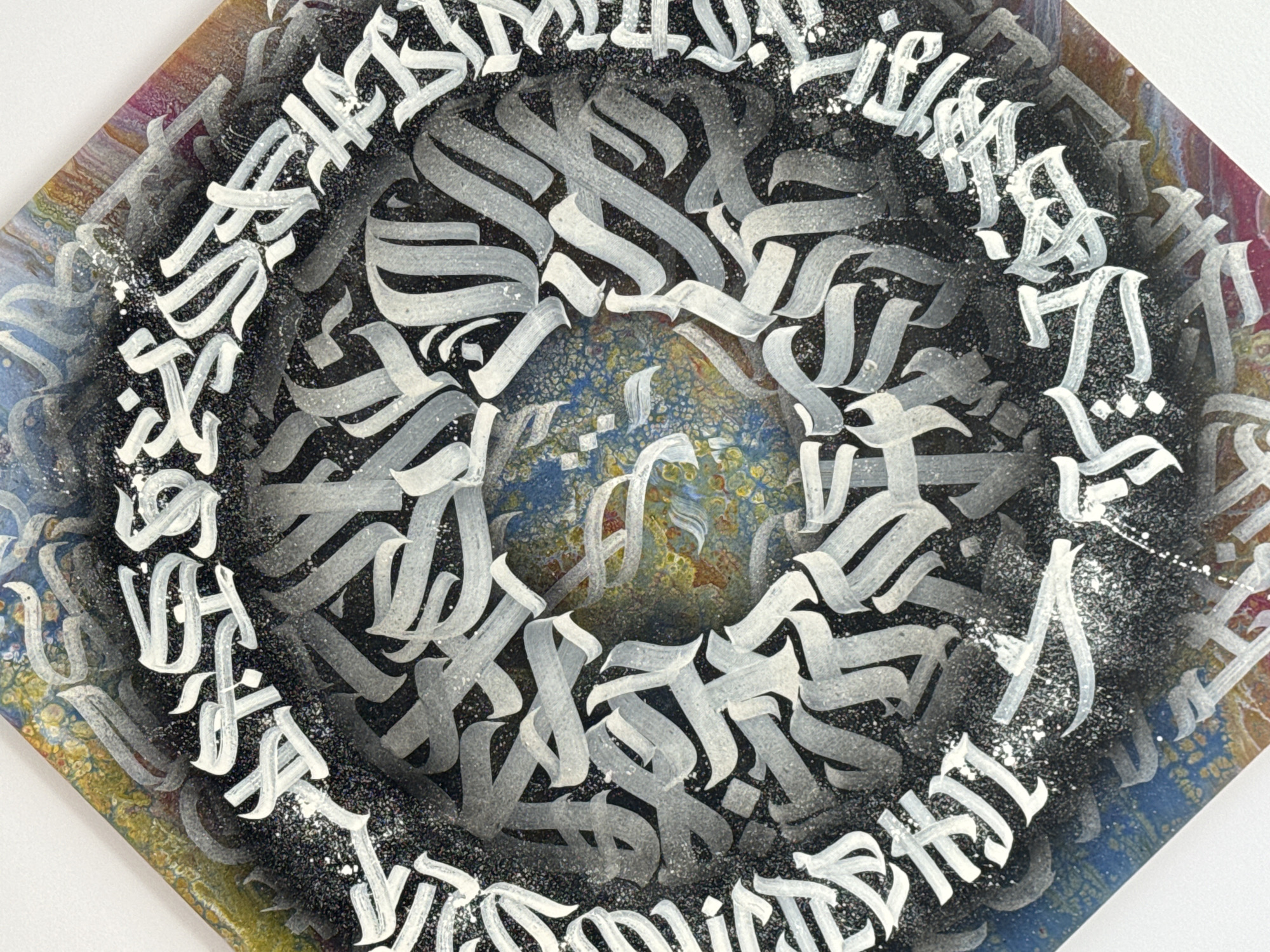 This is a close-up, top-down, vertical shot of a piece of abstract art. The art is on a diamond-shaped canvas, with a border of colorful paint that transitions from yellow and blue at the top left to pink and purple at the bottom right. The main composition features a central circular area of marbled blue and yellow paint, surrounded by black spray paint and white Arabic calligraphy. The calligraphy is intricate and layered, creating a sense of depth and movement. Some of the white calligraphy is speckled with white paint, resembling stars or a starry sky. The overall impression is one of dynamic energy and spiritual depth.