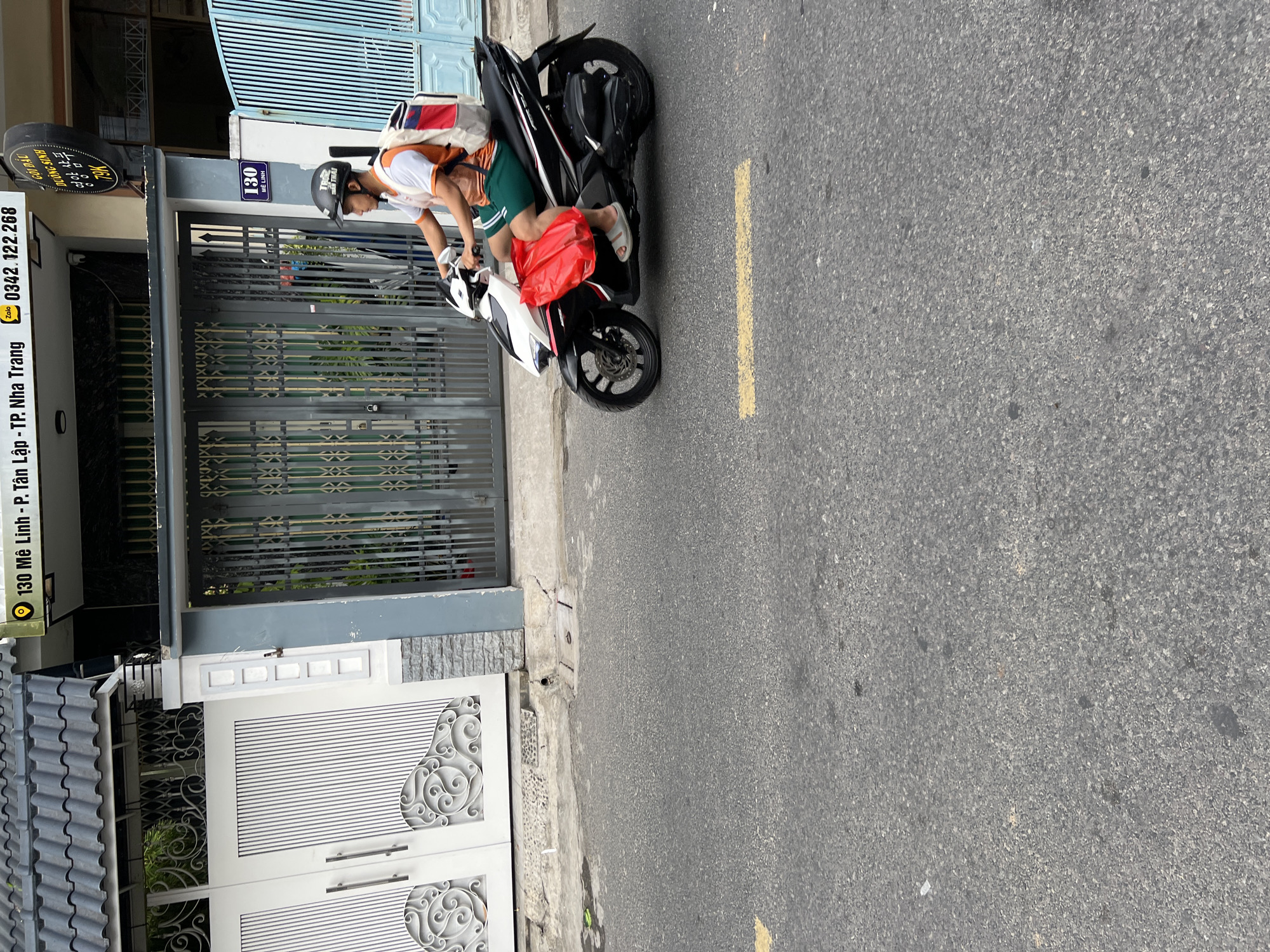 A man rides a black and white motorcycle down a paved street in Nha Trang, Vietnam. He is wearing a white t-shirt with orange trim, green shorts, a helmet, and sandals. A red plastic bag is attached to the front of the motorcycle. He is carrying a backpack with red and white accents. The street is bordered by buildings with decorative gates. A sign with Vietnamese writing is visible on one of the buildings.