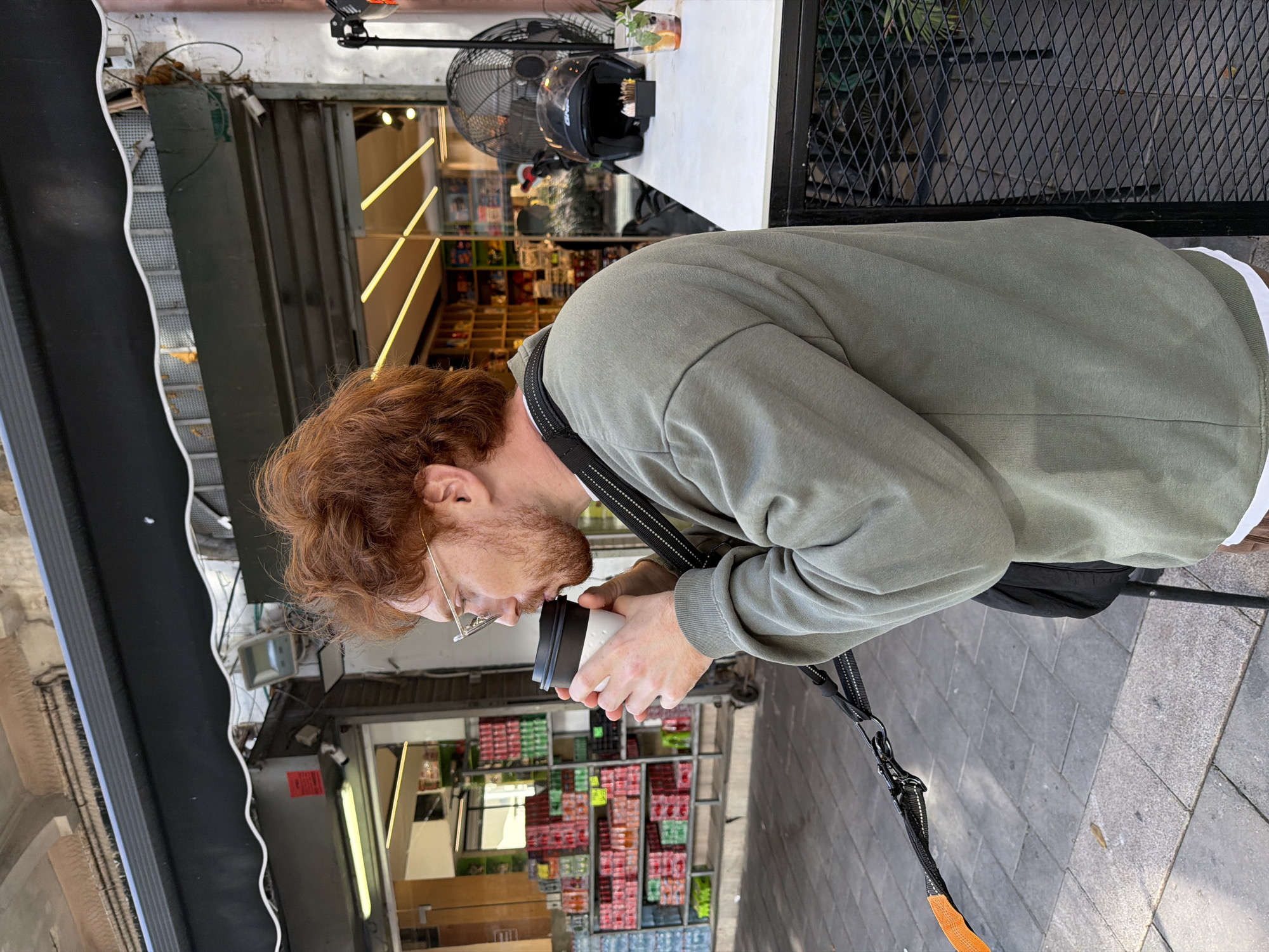 A man with reddish hair and glasses is drinking from a white paper cup with a black lid, standing on a sidewalk. He is wearing an olive green sweatshirt and a black strap is visible across his shoulder. The sidewalk is paved with gray bricks. Behind him, there is a store with shelves filled with various products. A black awning is over the front of the store. Reflected in the window are linear light fixtures. On a nearby table, there is a black motorcycle helmet and a black fan. The location is likely in Tel Aviv, Israel, considering the architectural style and the Hebrew text on the store sign.